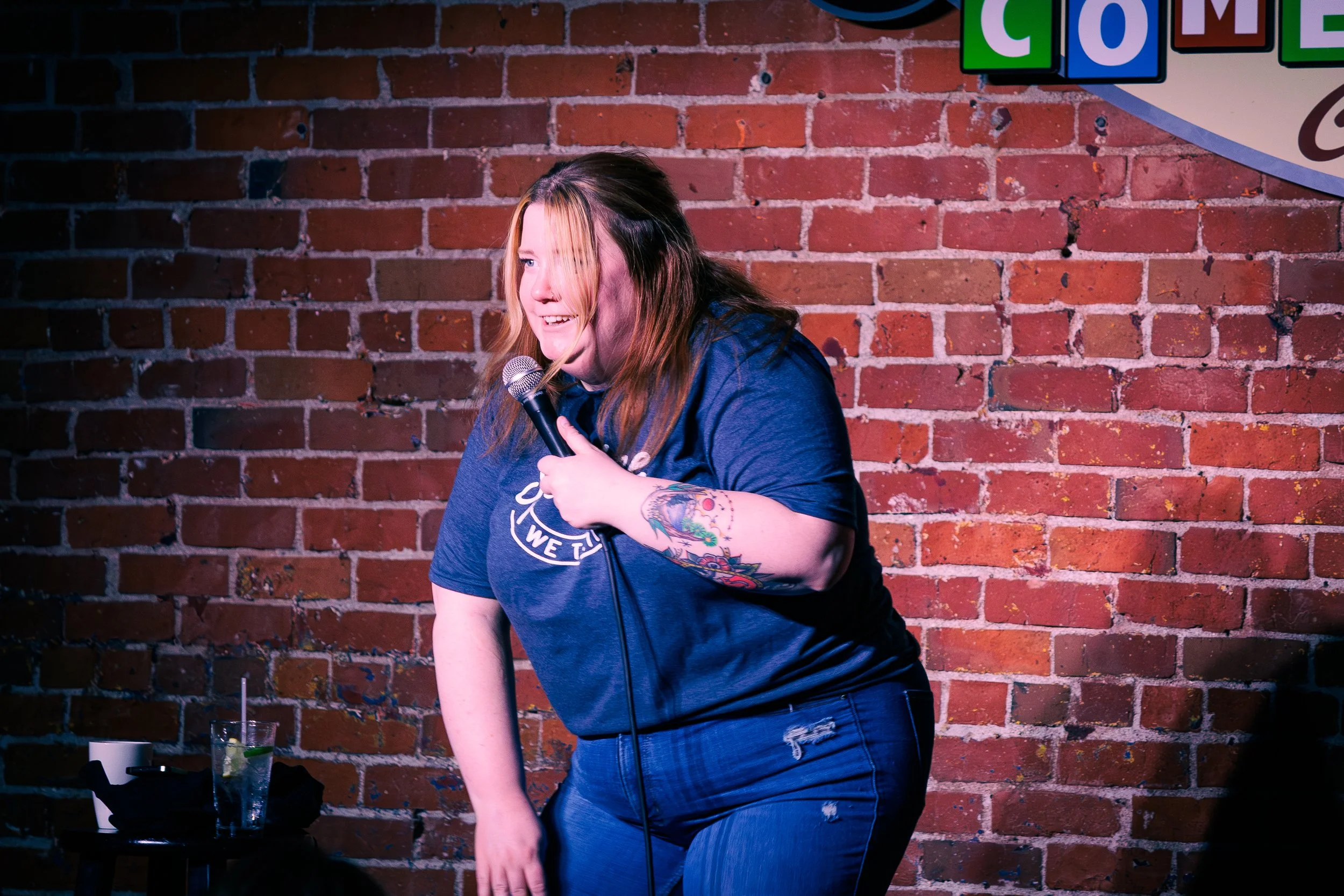 A woman with long hair and tattoos on her arm is holding a microphone while performing comedy on stage against a brick wall background.