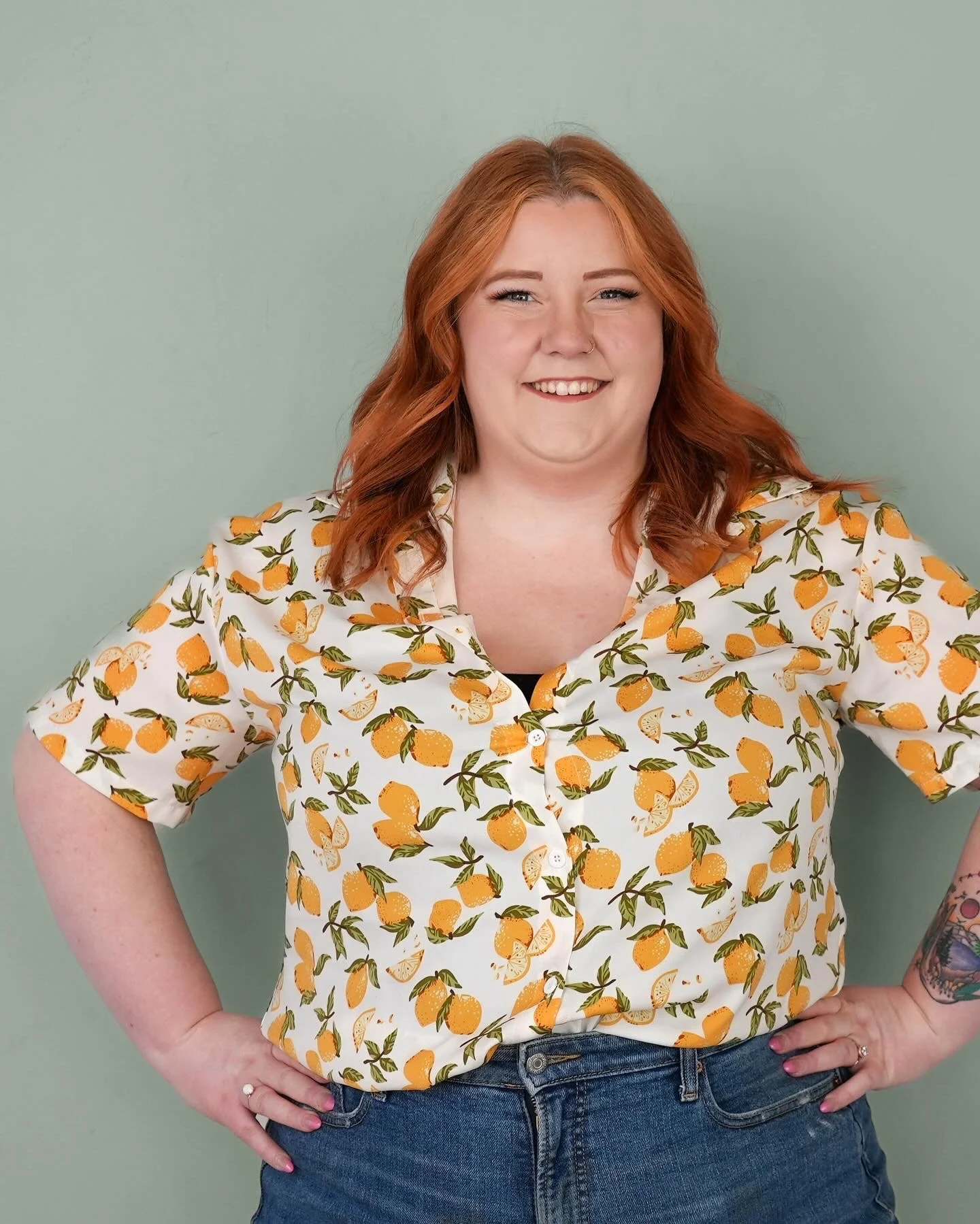A woman with red hair smiling, wearing a white shirt with a lemon print and blue jeans, standing against a light green background.