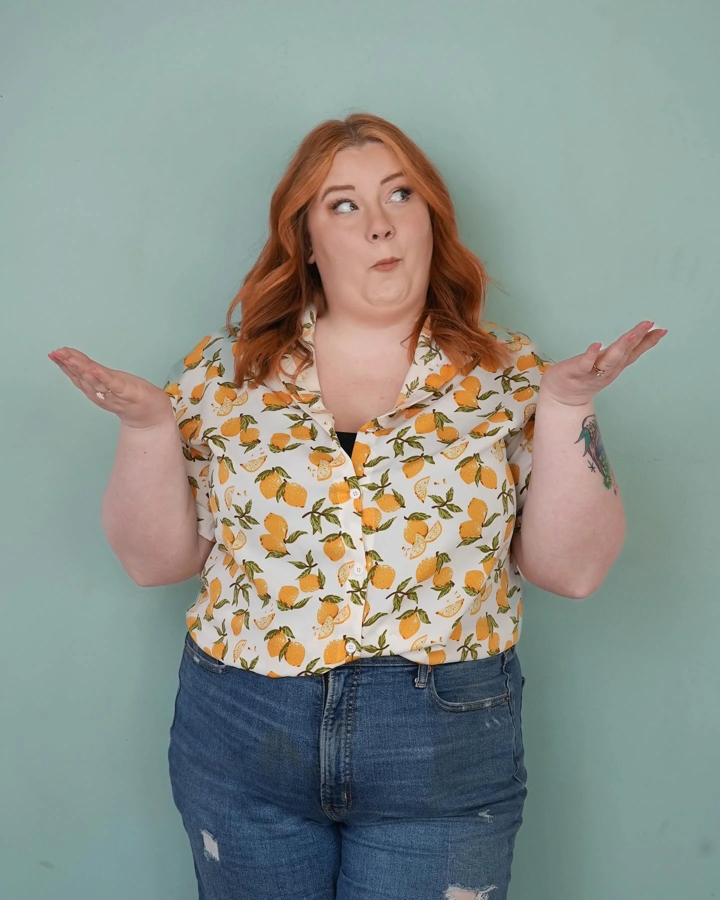 A woman with red hair and tattoos on her arms stands against a plain pastel green background, wearing a short-sleeved button-up shirt with a lemon pattern and distressed jeans. She has her arms raised, palms facing up, with a puzzled or questioning e