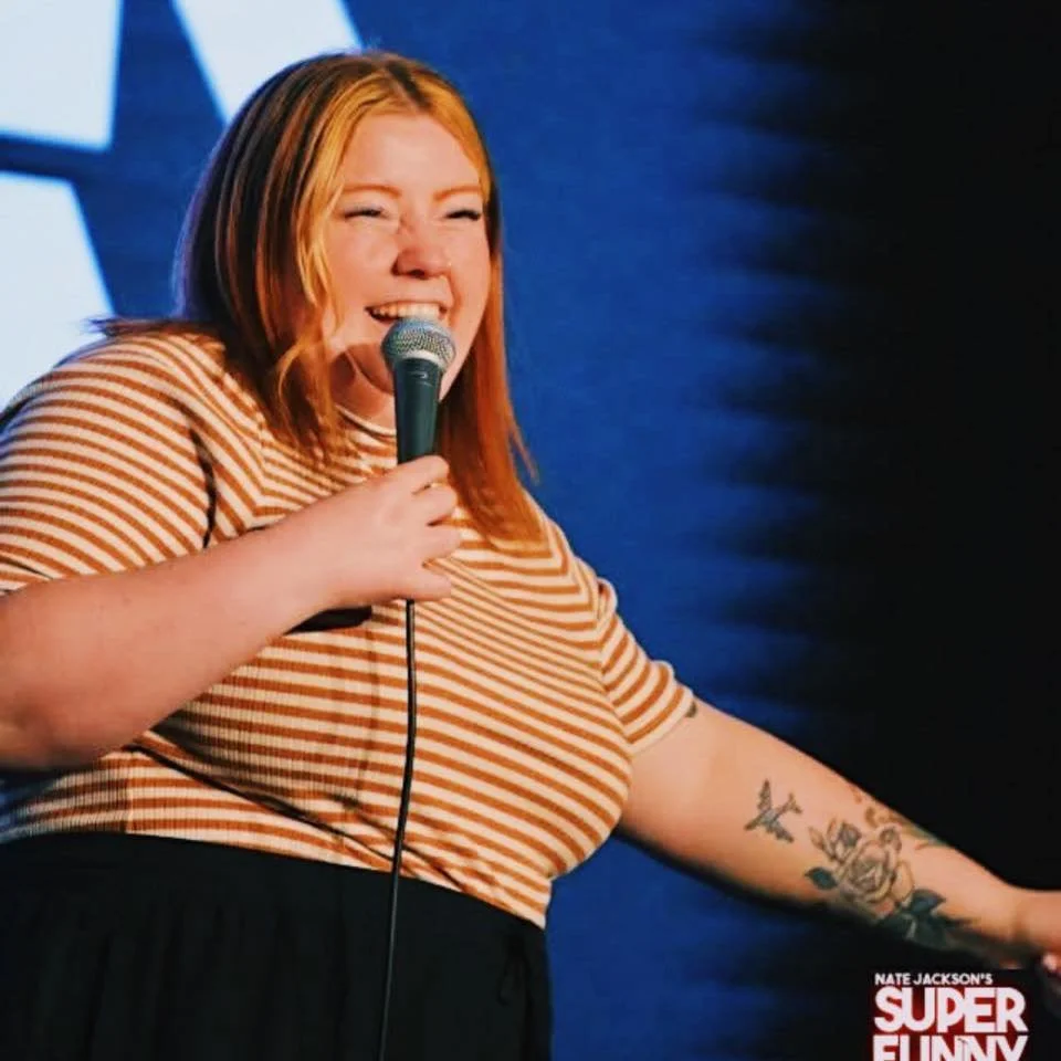 A woman with red hair wearing a striped shirt holds a microphone and is laughing. She has tattoos on her left forearm, and the background is dark with stage lighting.