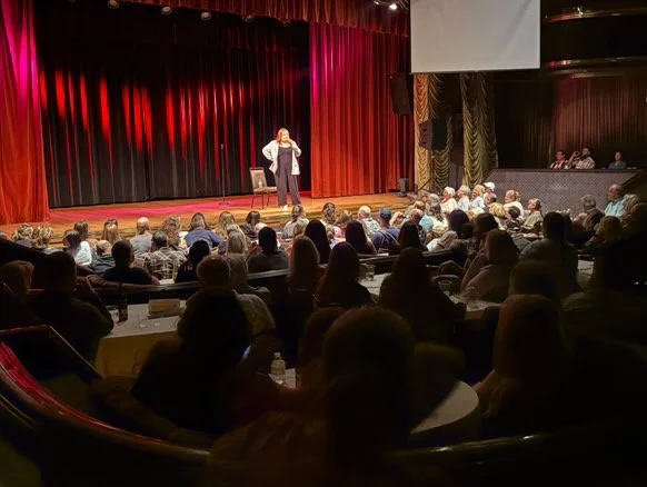A woman performs on stage in front of a large audience in a theater with red curtains and a dark background, with some people seated at tables in the audience.