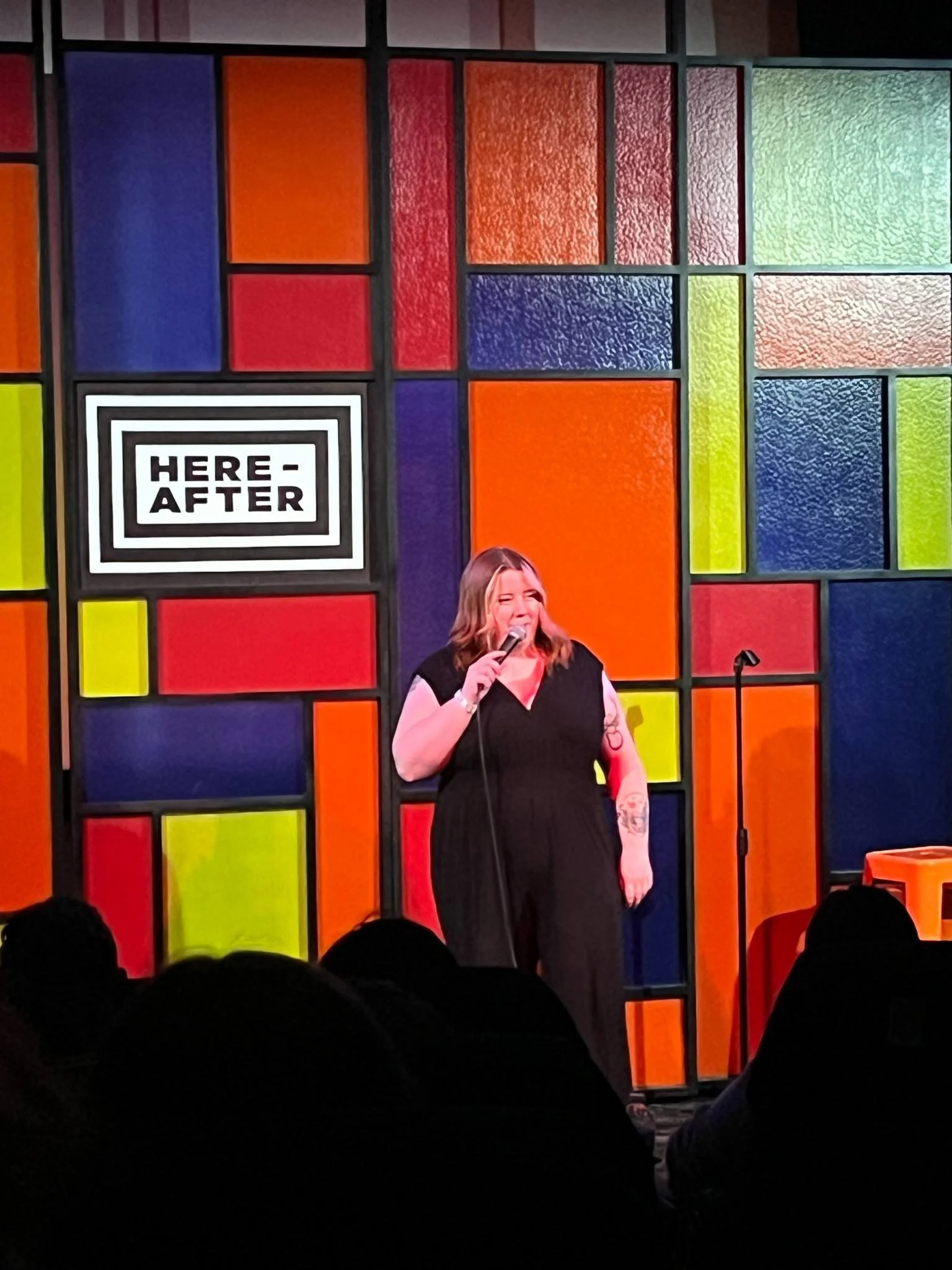 A woman with blonde hair and tattoos, wearing a black outfit, holding a microphone and performing on stage at the Here After comedy club, with a colorful geometric background.