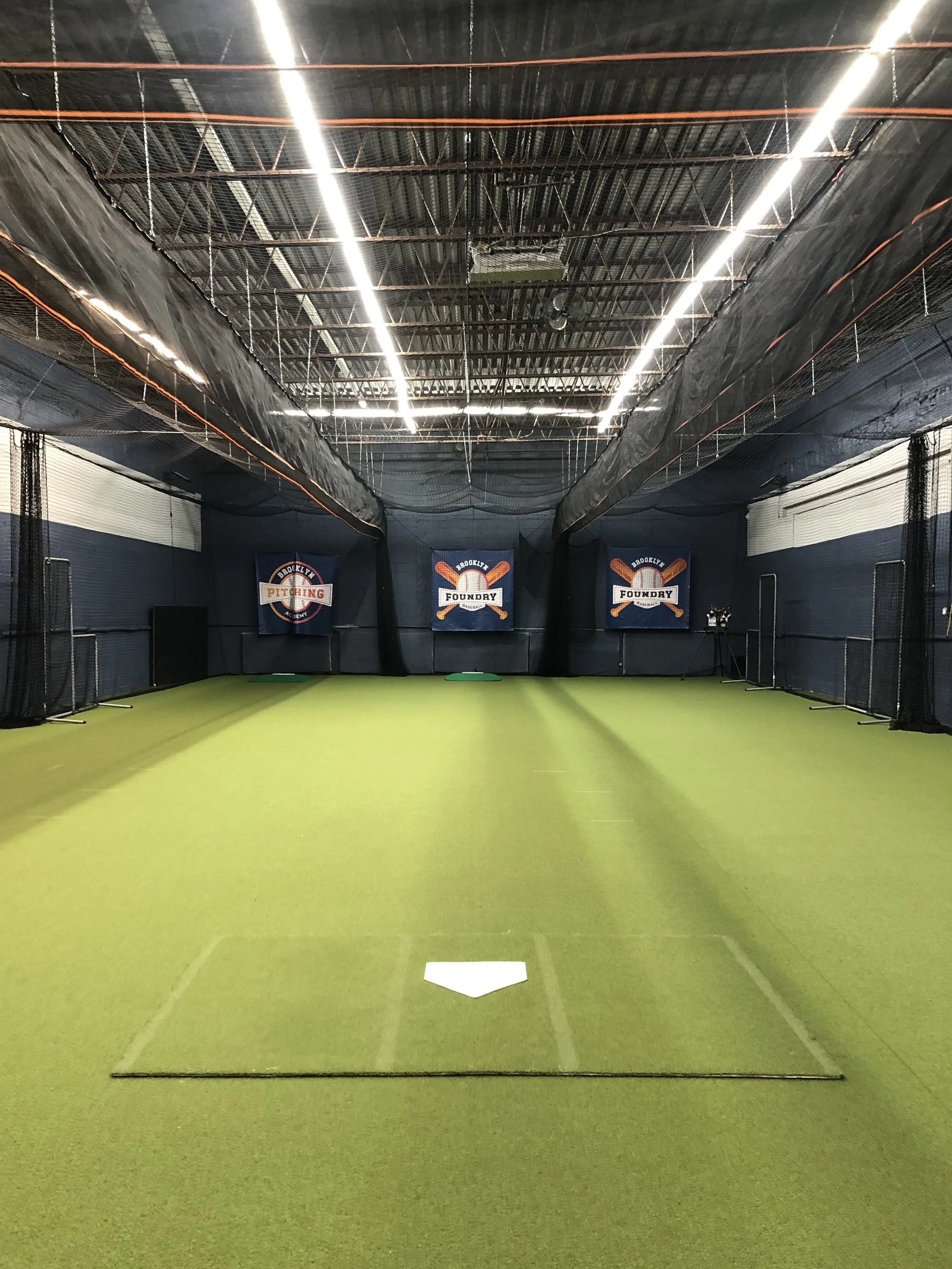 Indoor baseball or softball practice area with a green turf field, pitching mound, and backstop banners for Brooklyn Pithing and Foundry.