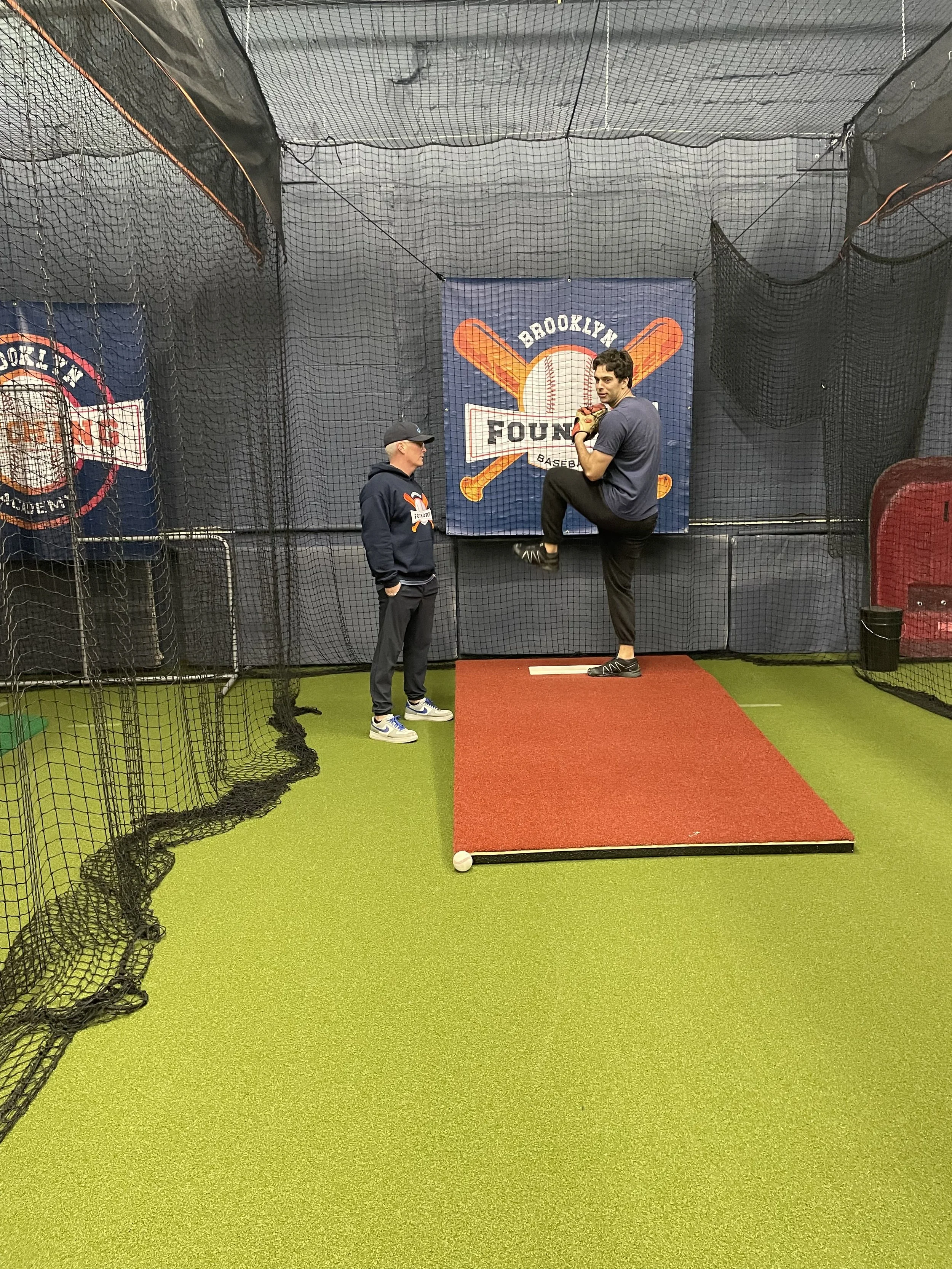 Two men in an indoor batting cage at Brooklyn Baseball Academy, one practicing pitching with cleats and gloves, the other observing. You can see netting, a sign, and artificial turf.