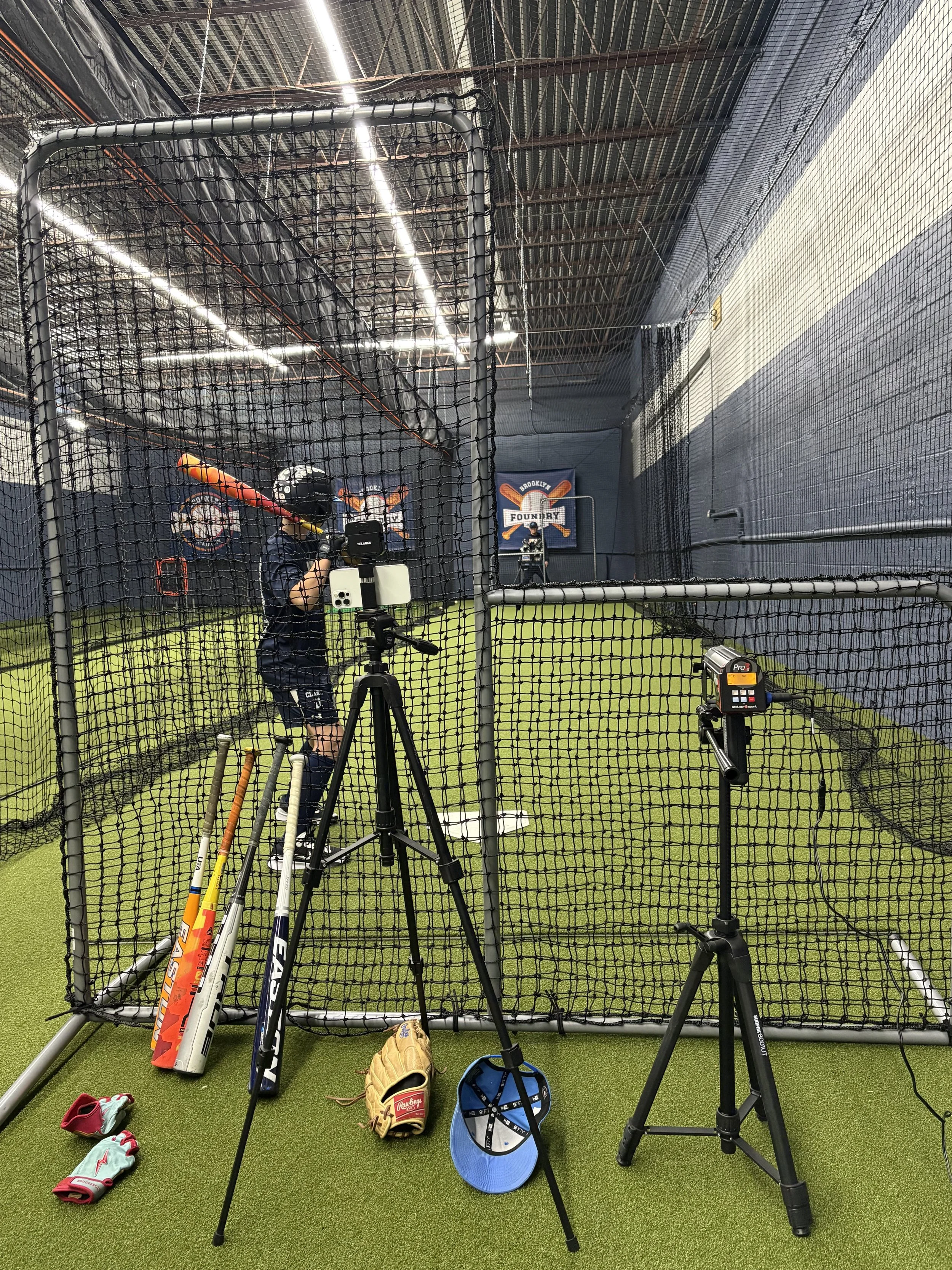 Indoor baseball or softball batting practice area with a batter inside the cage holding a bat. Equipment including helmets, bats, a glove, and pitch tracking devices are visible in the foreground. The background features a banner with a baseball theme and a regulation-sized batting cage.
