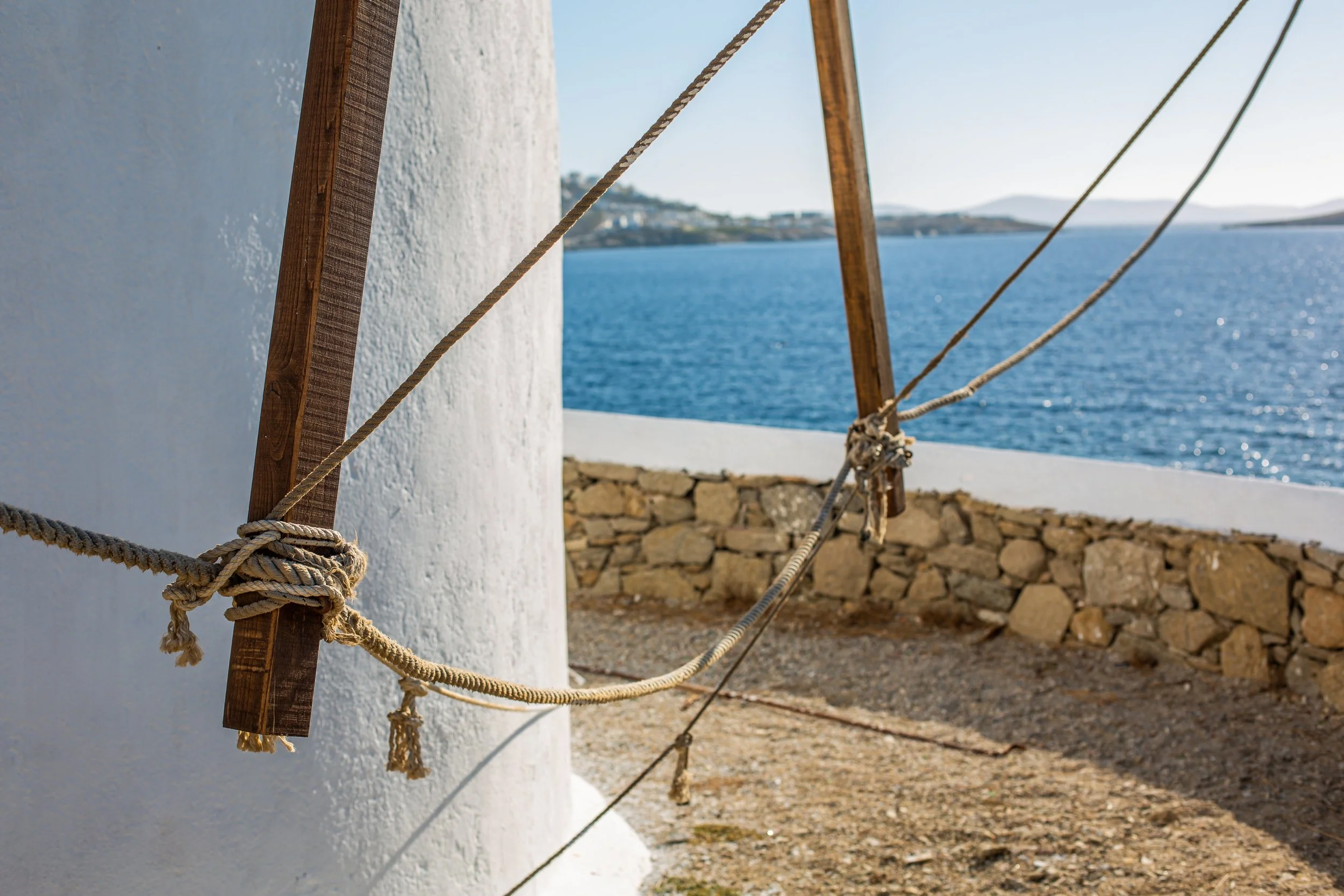 Rope tied around a wooden support structure against a white wall near a stone wall and blue water in the background.