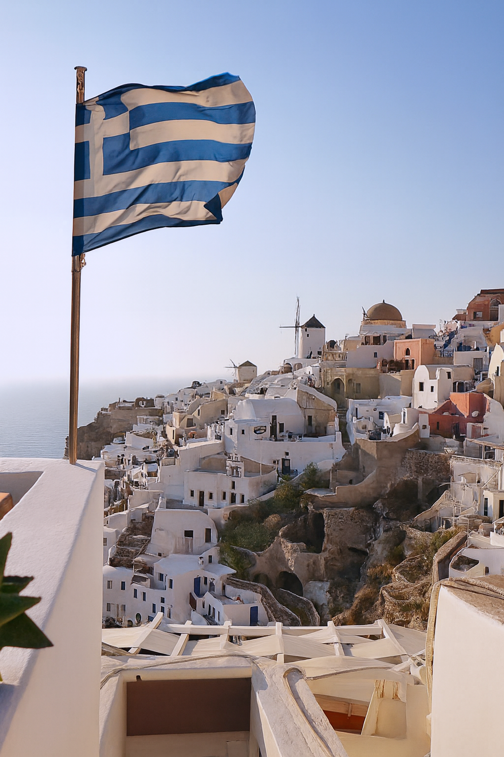 Greece flag flying over white buildings on a hillside with windmills, overlooking the sea, in Santorini.