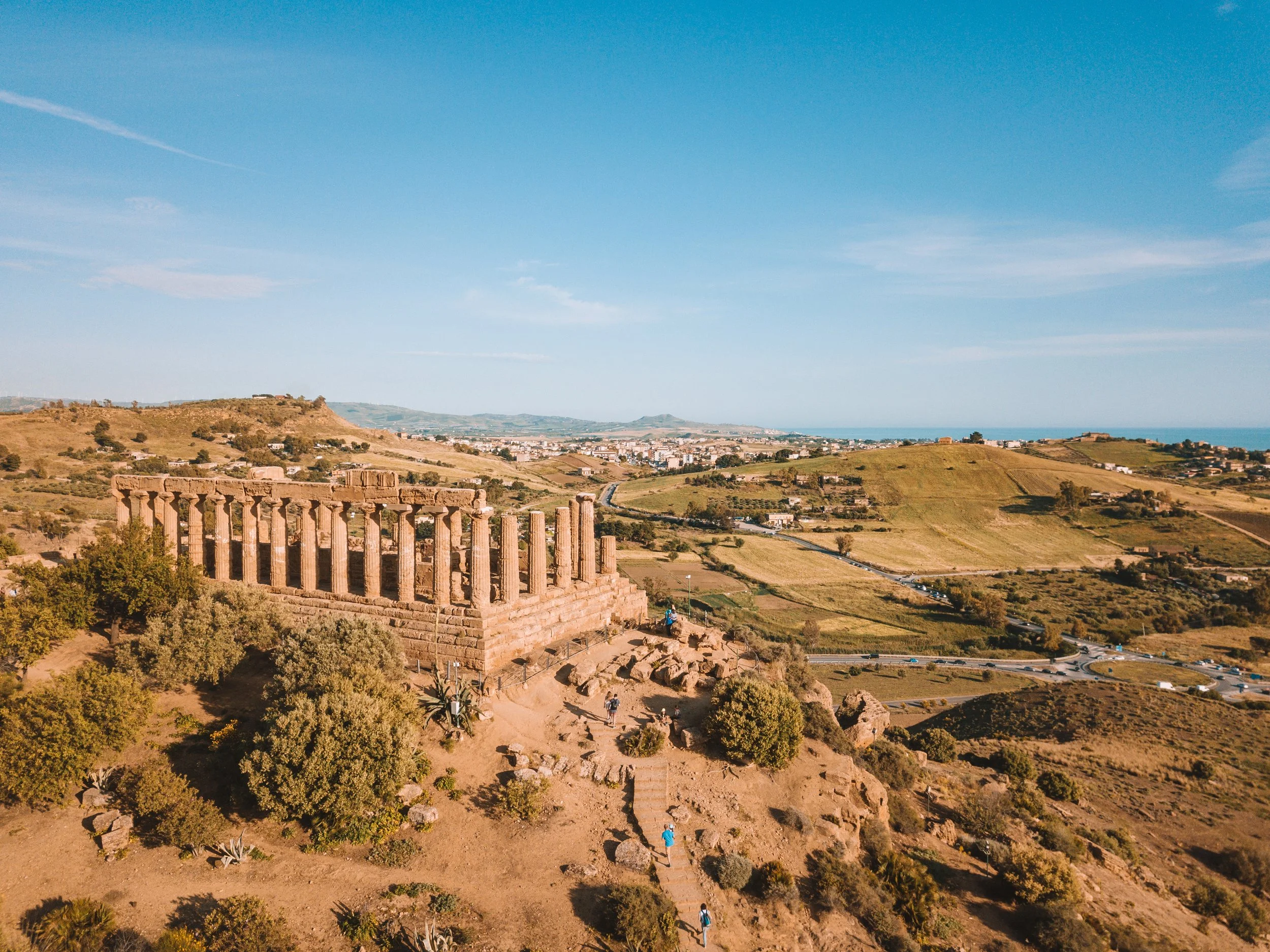 Ancient Greek ruins on a hill overlooking a landscape with rolling hills, a town in the distance, and a blue sky with some clouds.
