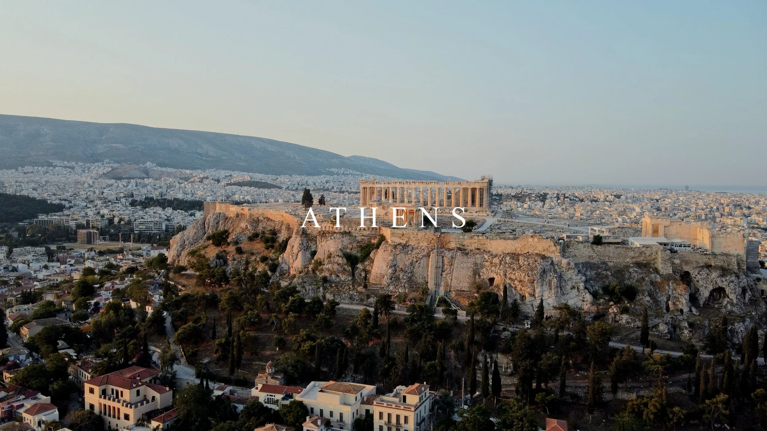 View of the Acropolis and Parthenon in Athens, Greece, during sunset, with the cityscape and mountains in the background.