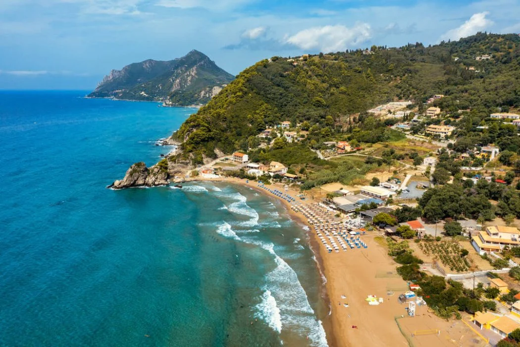 Aerial view of a beach with sun umbrellas, waves, and a hillside with houses and greenery