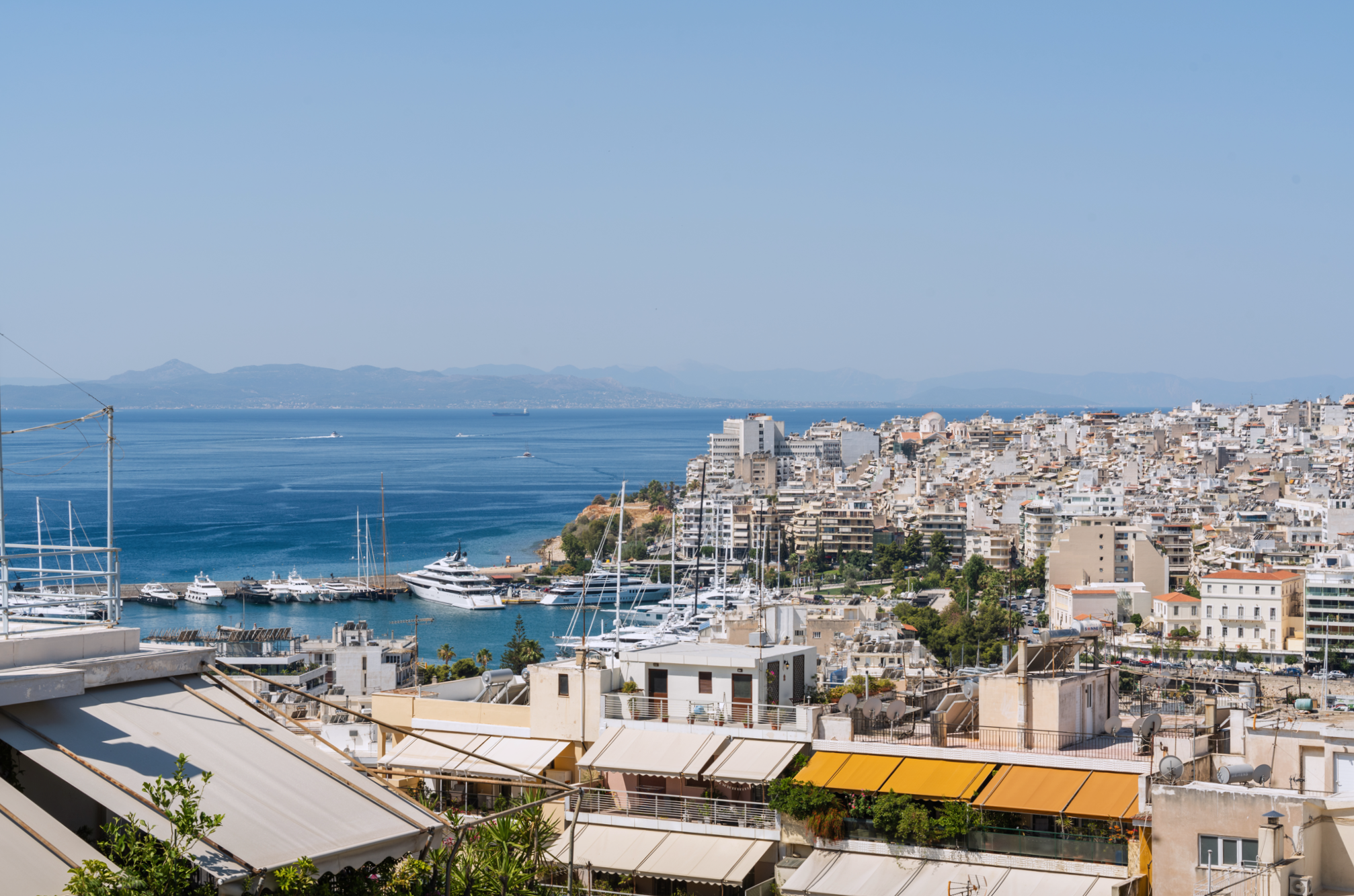 A harbor with yachts and boats, surrounded by white buildings, with a cityscape and mountains in the background under a clear blue sky.