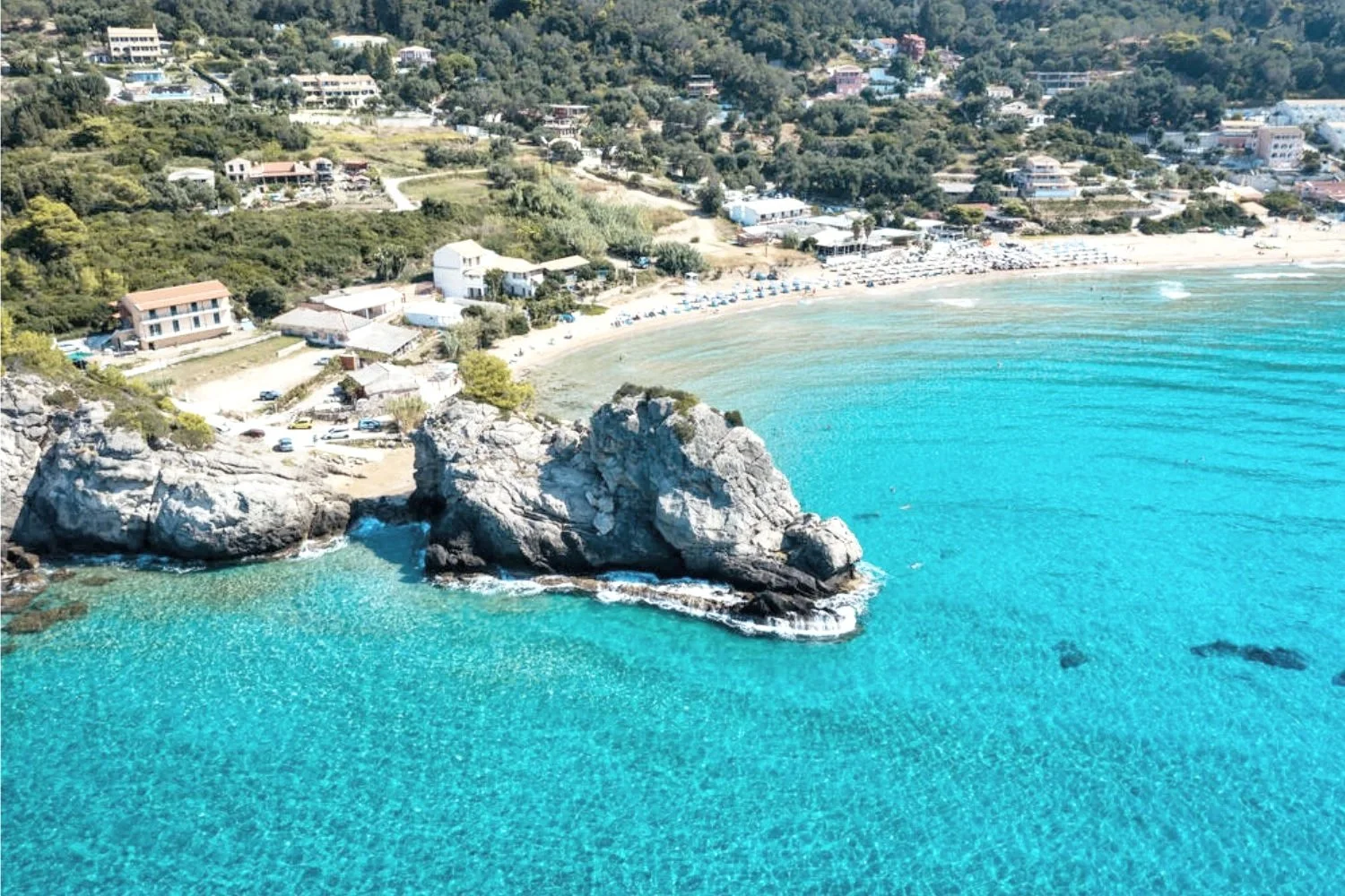 Aerial view of a coastal beach with turquoise water, a rock formation extending into the sea, and a shoreline with umbrellas and sunbathers. Residential houses and greenery are on the hillside behind the beach.