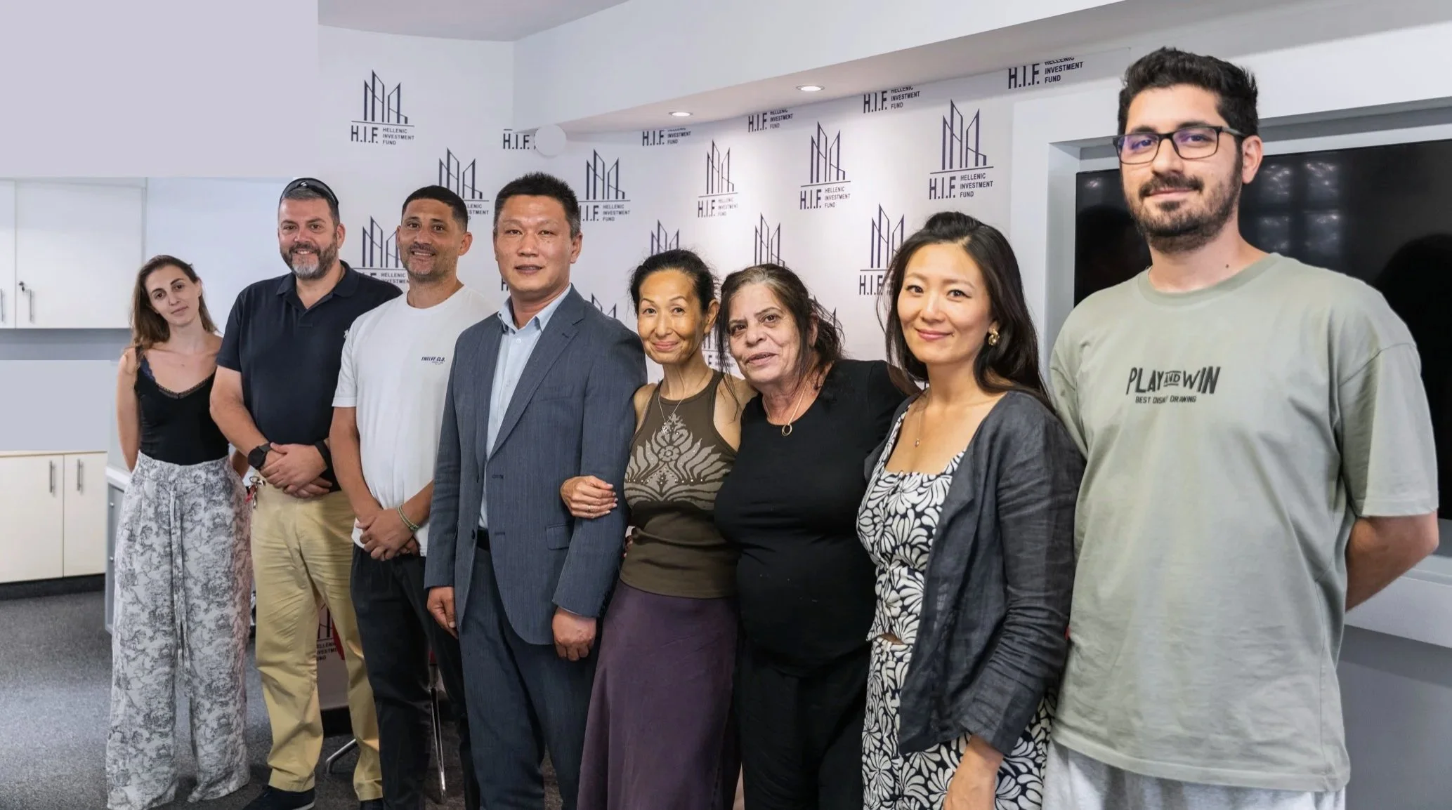 Group of nine diverse individuals standing side by side in an office or conference room with a backdrop that reads 'Hellenic Investment Fund'.