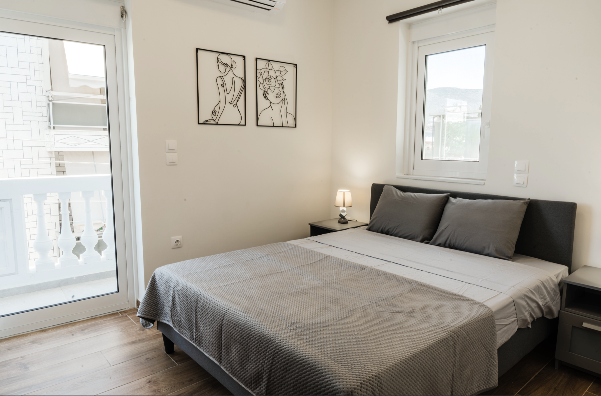 Minimalist bedroom with a bed, grey pillows, nightstands, a small lamp, wall art of women, window, and balcony door.