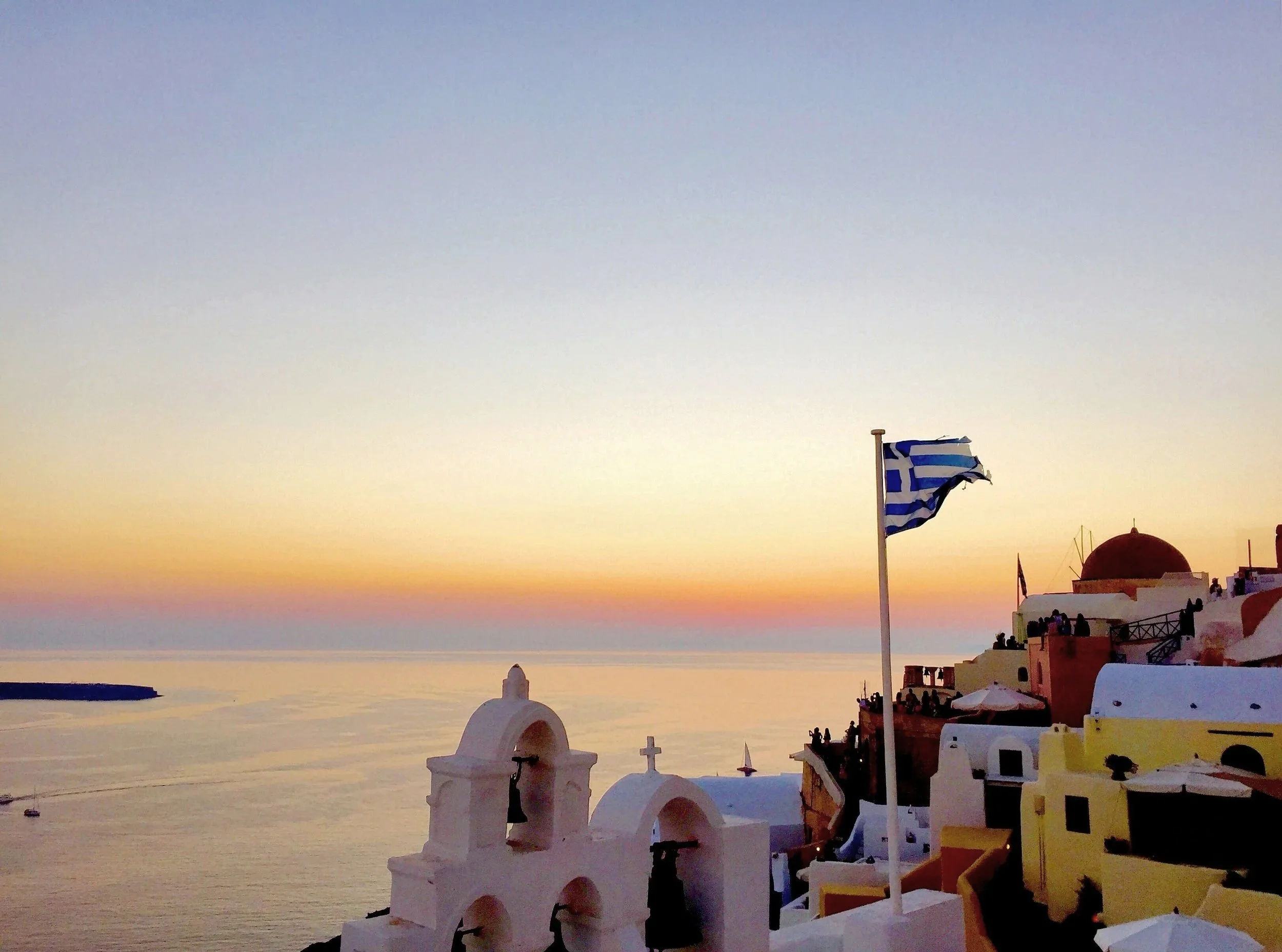 Greek flag flying over a seaside village at sunset with white buildings and domed roofs.
