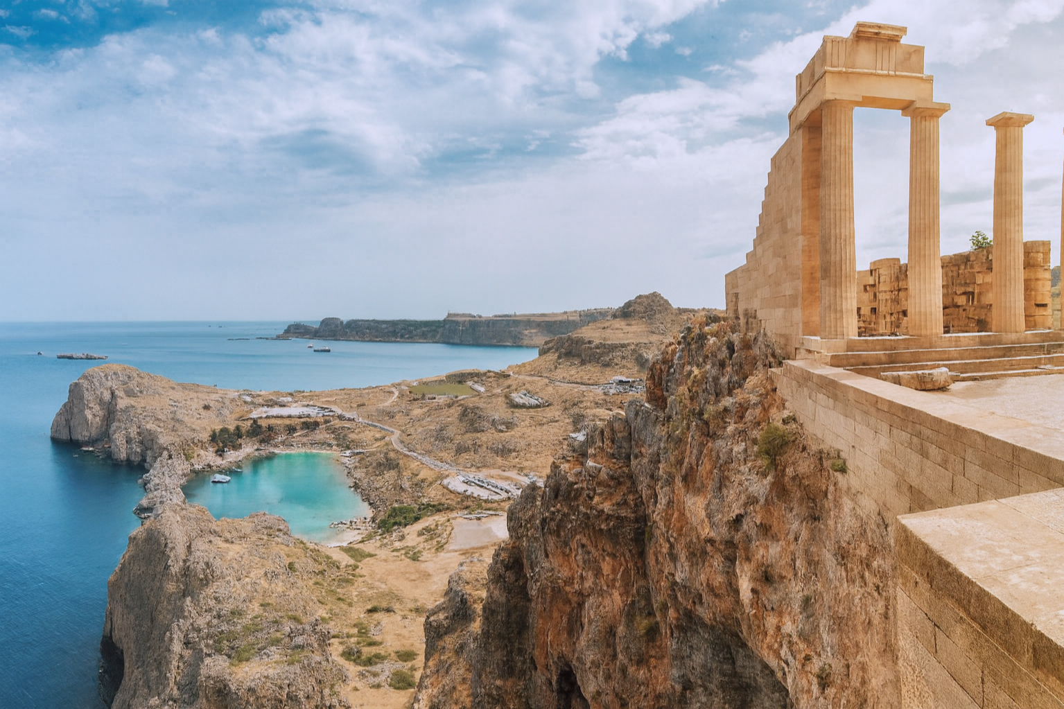 Ancient Greek ruins on a cliff overlooking the sea, with a blue sky and scattered clouds.