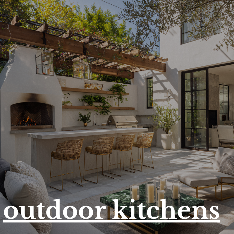 Outdoor kitchen with white stucco fireplace, built-in grill, wooden shelves with plants, wicker barstools, large potted plant, and seating area with candles, surrounded by trees and modern house with large windows.