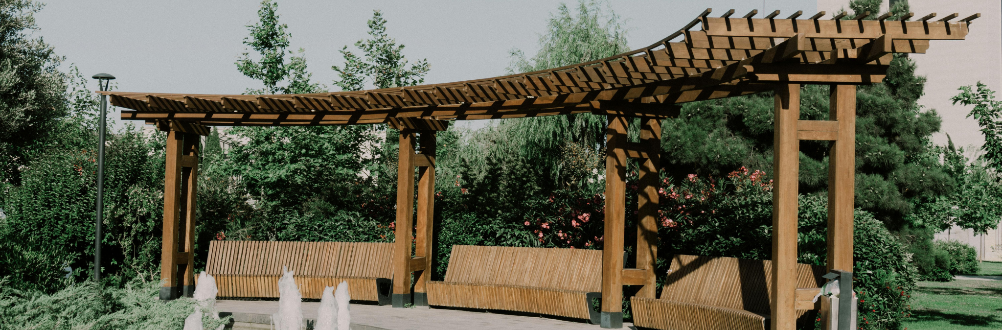 Outdoor wooden shelter with curved roof, two benches underneath, surrounded by lush greenery.
