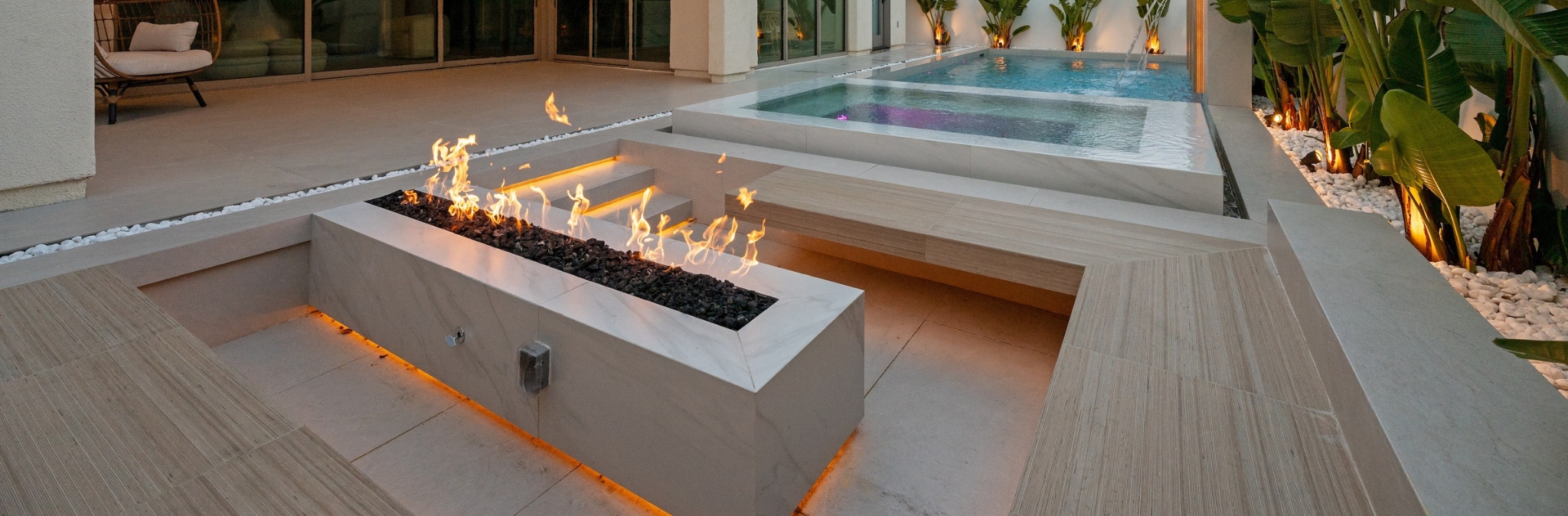 Modern outdoor pool area with hot tubs, a fire feature, and lush green plants at dusk.