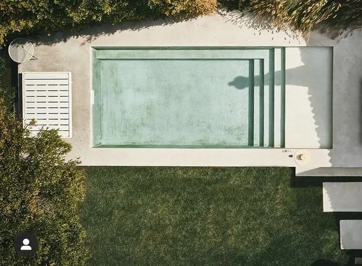 Bird's eye view of a rectangular swimming pool with steps, surrounded by a concrete patio and green grass, with trees and bushes along one side.
