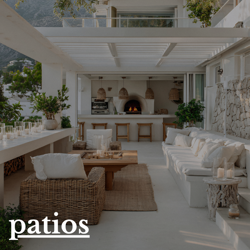 Outdoor patio area with a white sofa, wicker chair, wooden coffee table, bar stools, built-in kitchen with pizza oven, and string lights in a sunny, modern setting.