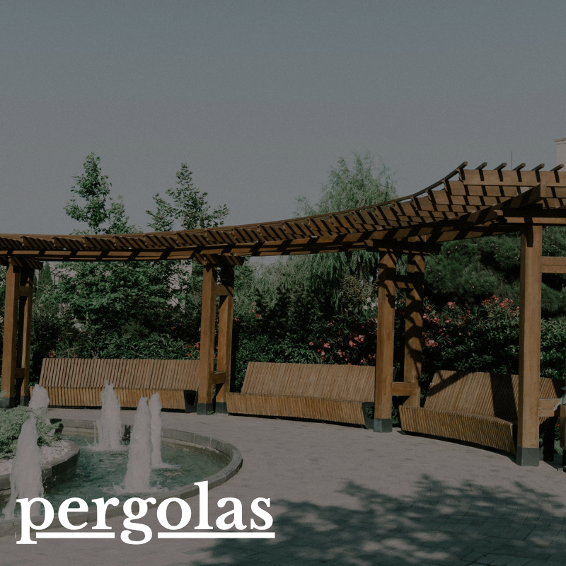 Wooden pergola with curved roof, three water fountains in foreground, wooden benches along the structure, lush green trees and bushes in the background.