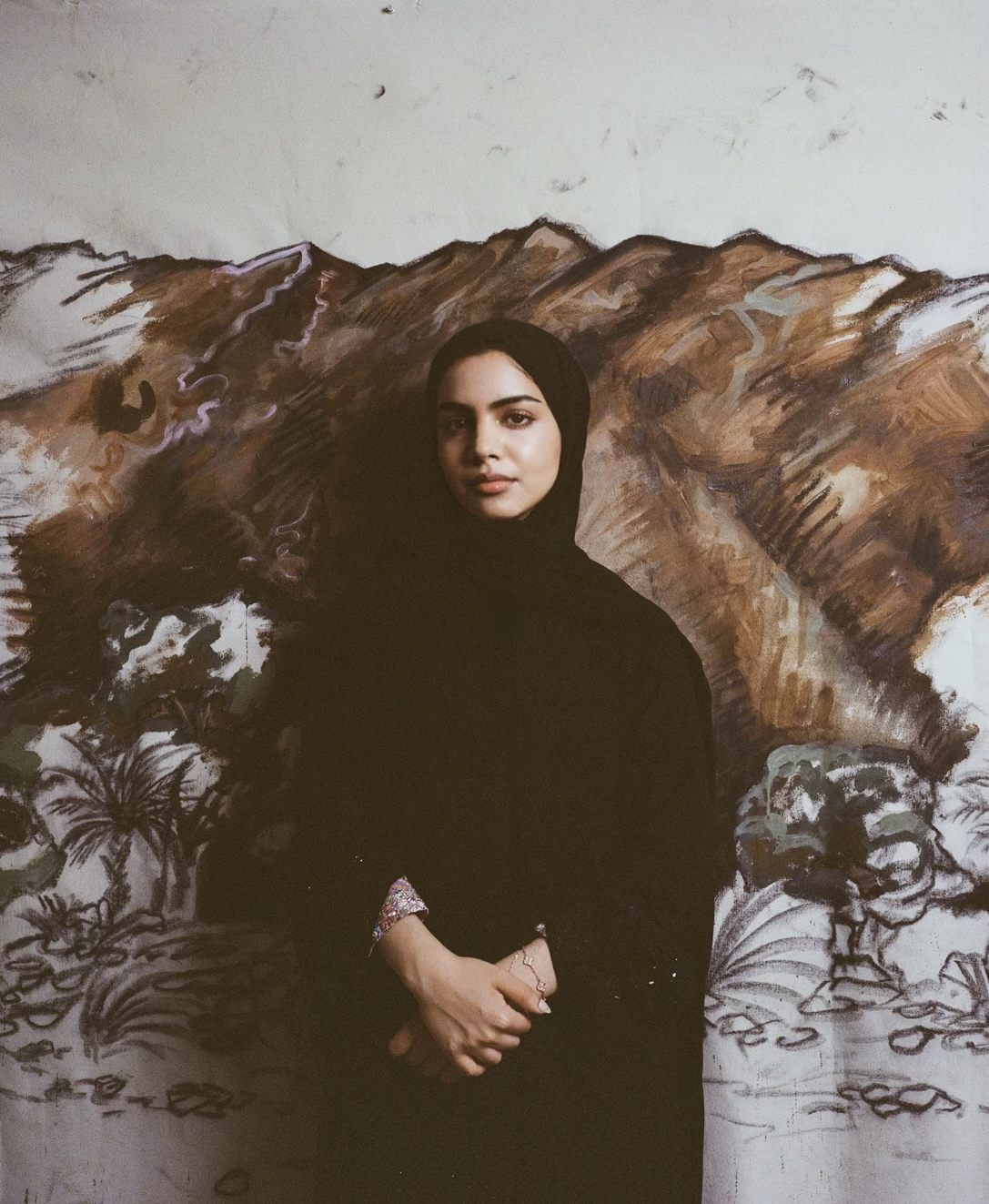 A woman wearing a black hijab and dark clothing standing in front of a mural of mountainous landscape and trees painted on a wall.