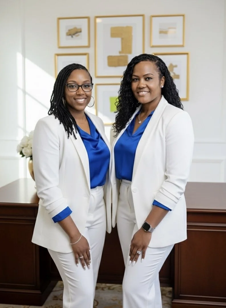 Two women dressed in white suits and blue blouses, standing in an office with framed abstract art on the wall behind them.