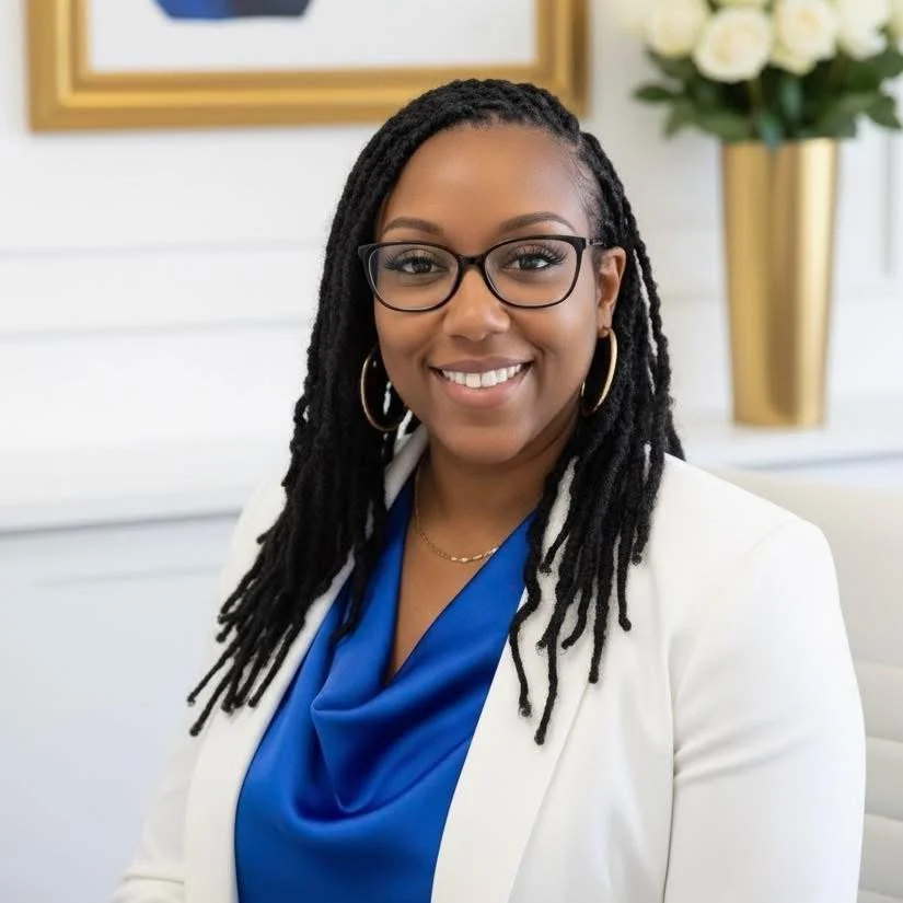 A smiling woman with glasses, wearing a white blazer and a blue top, seated in a bright, modern room with white walls, a framed picture, and a tall vase with white flowers in the background.