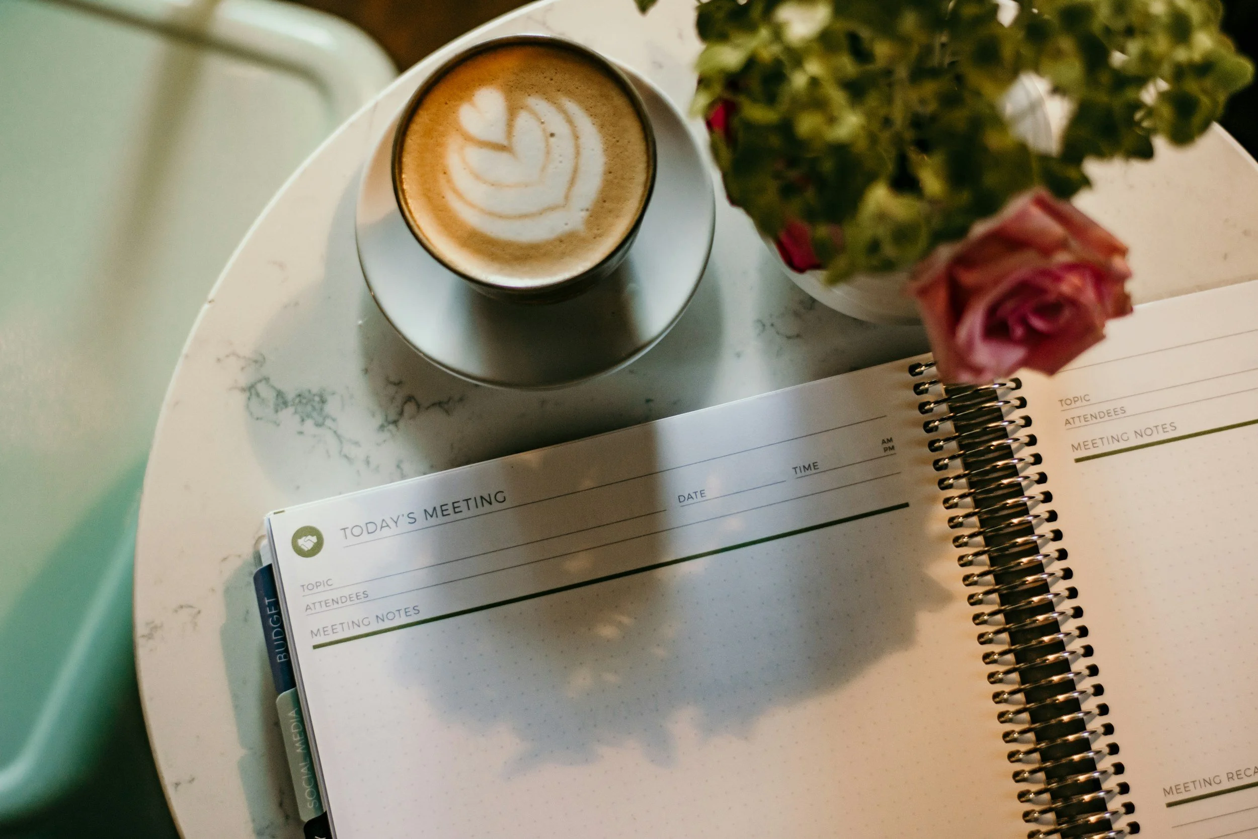A spiral notebook, a coffee cup with latte art, and a potted pink rose on a white marble table.