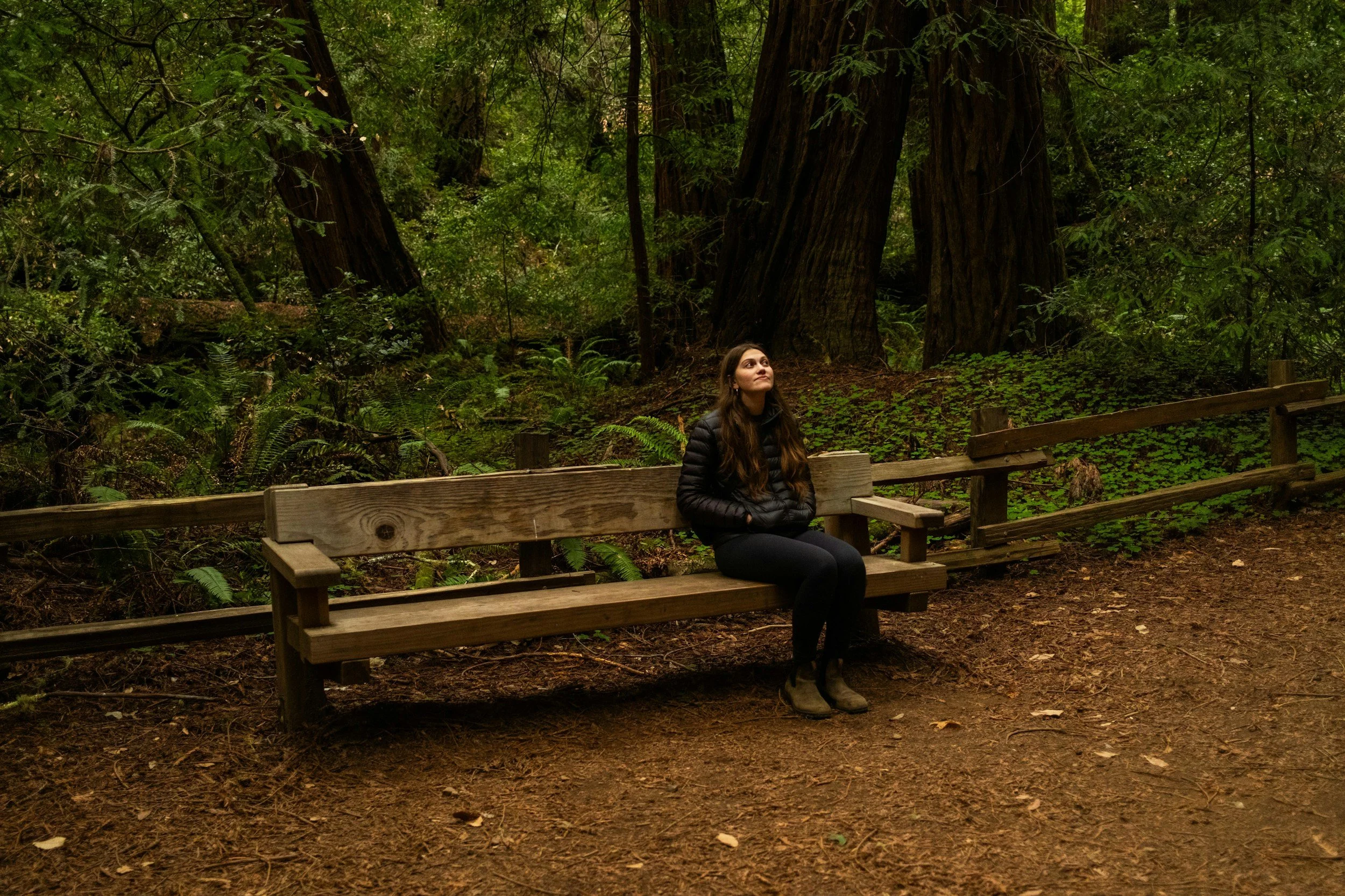 A woman sitting alone on a wooden park bench in a lush, green forest, looking upward.