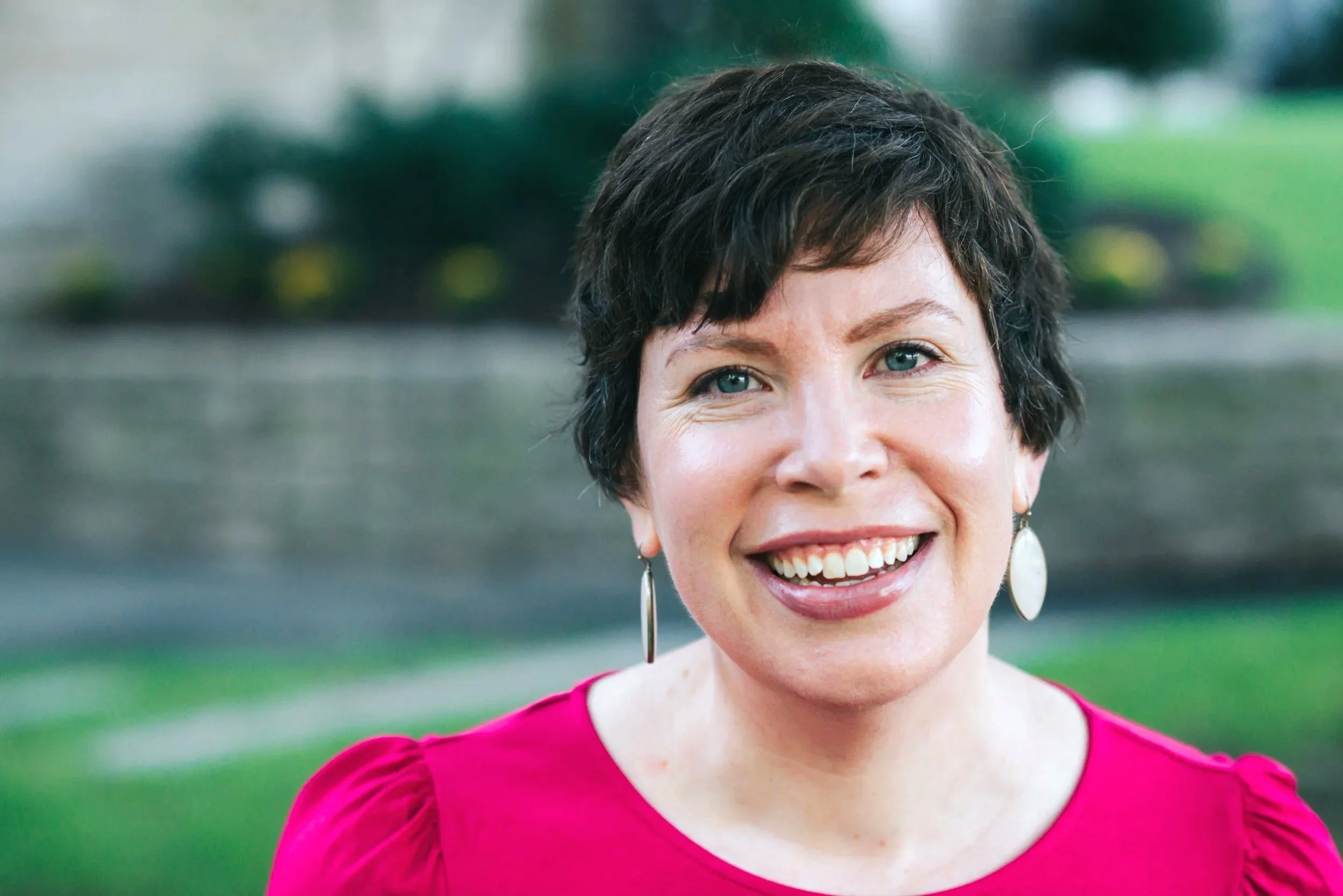 A woman with short dark hair and blue eyes smiling outdoors, wearing a pink top and white earrings.