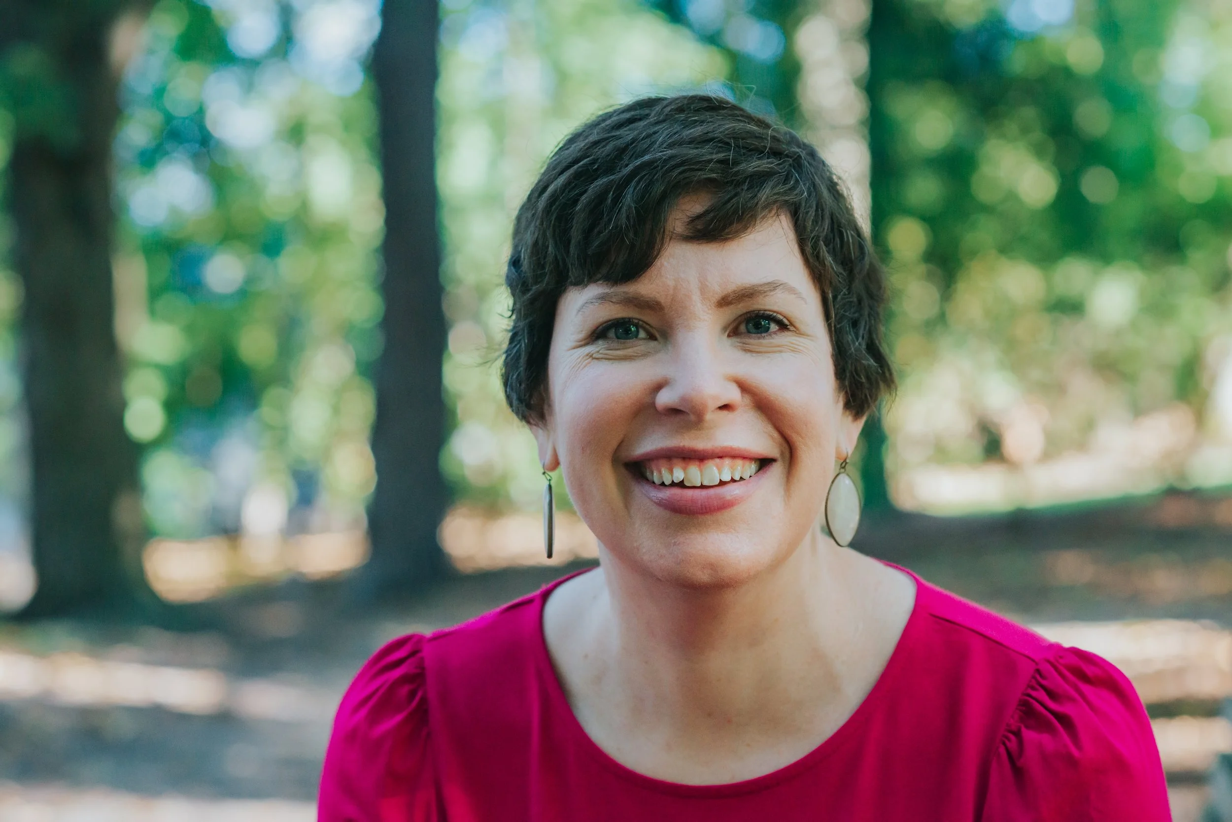 Smiling woman with short dark hair, wearing earrings and a pink top, outdoors with trees in the background.