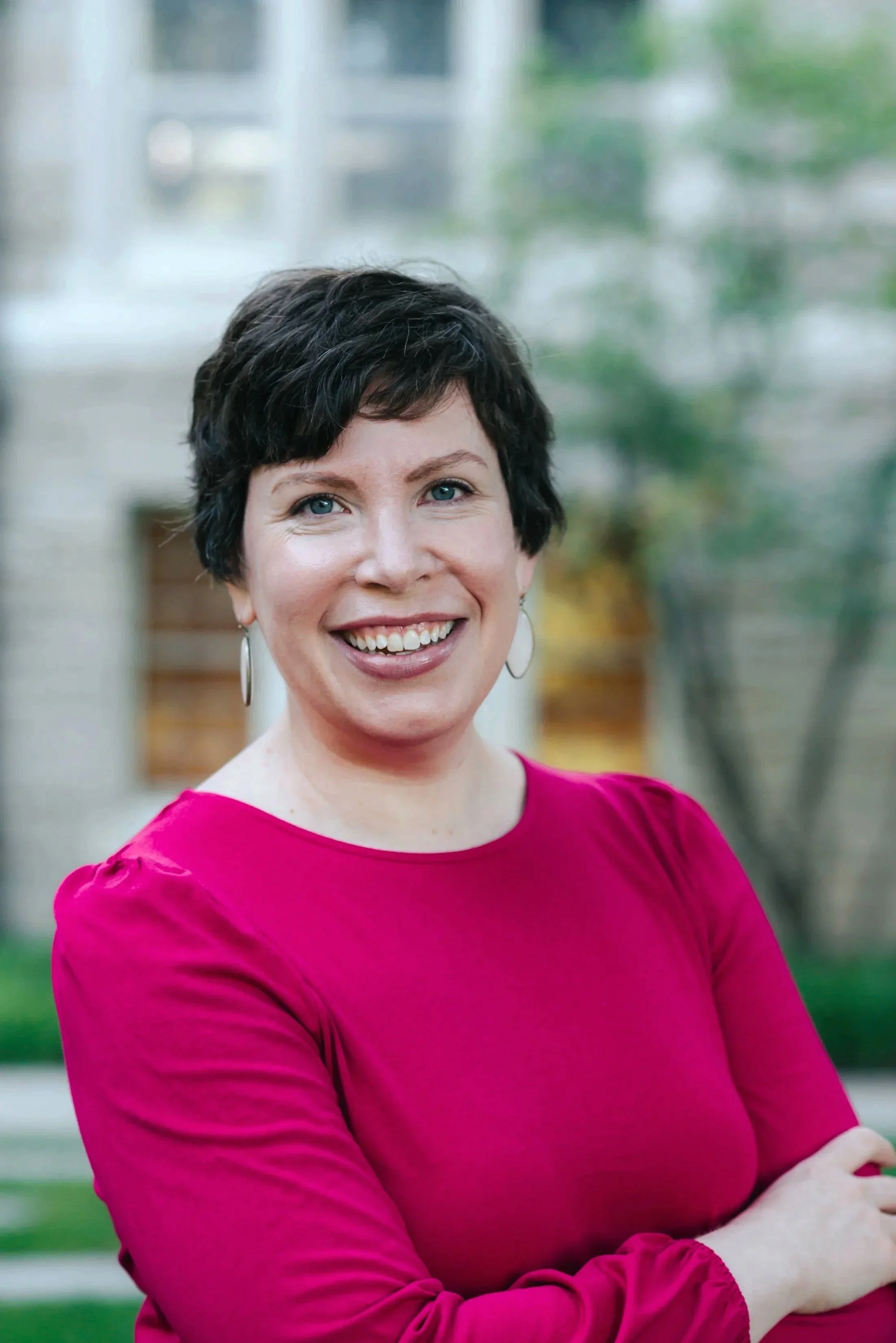 A woman with short dark hair, wearing a bright pink top, smiling outdoors.