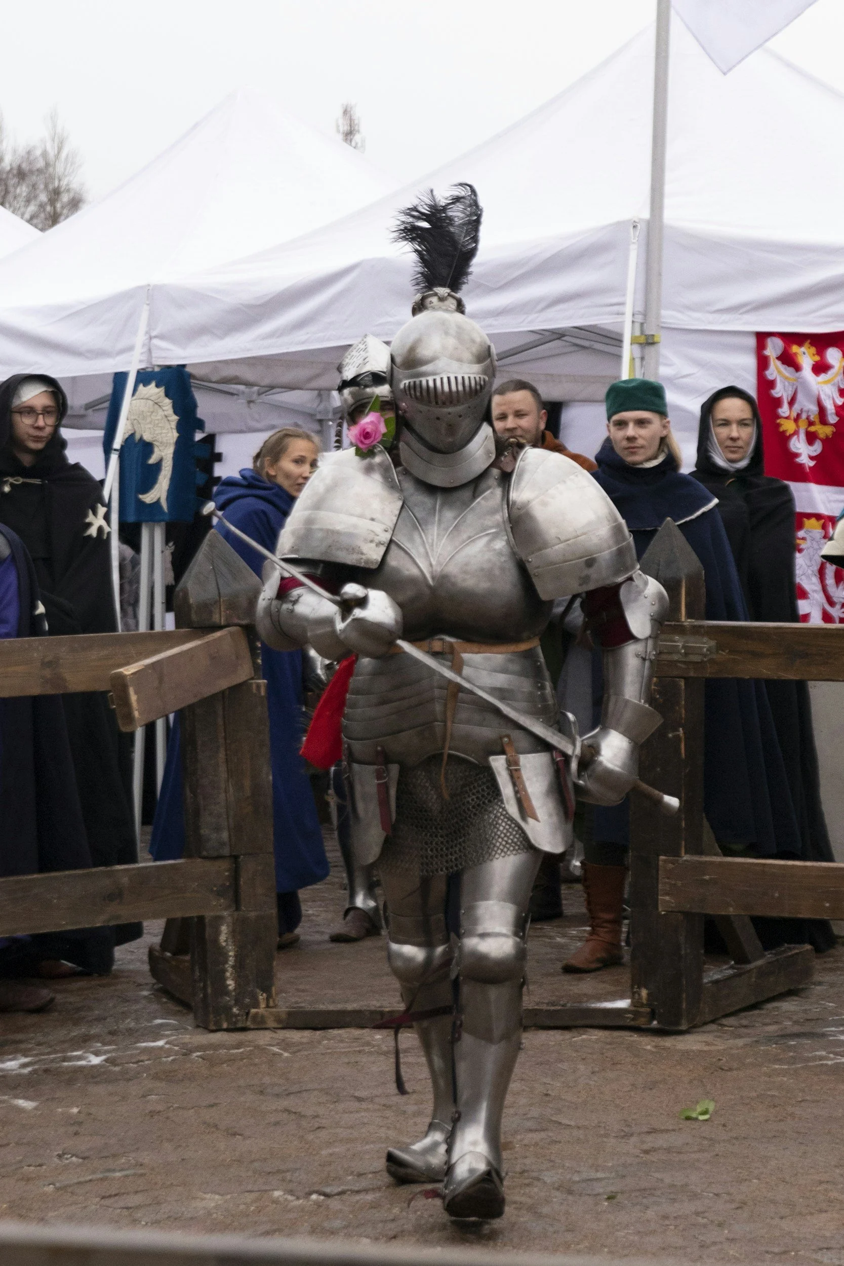 A person dressed in full medieval knight armor, holding a sword, walking in front of a group of people dressed in medieval costumes at a historical reenactment event.