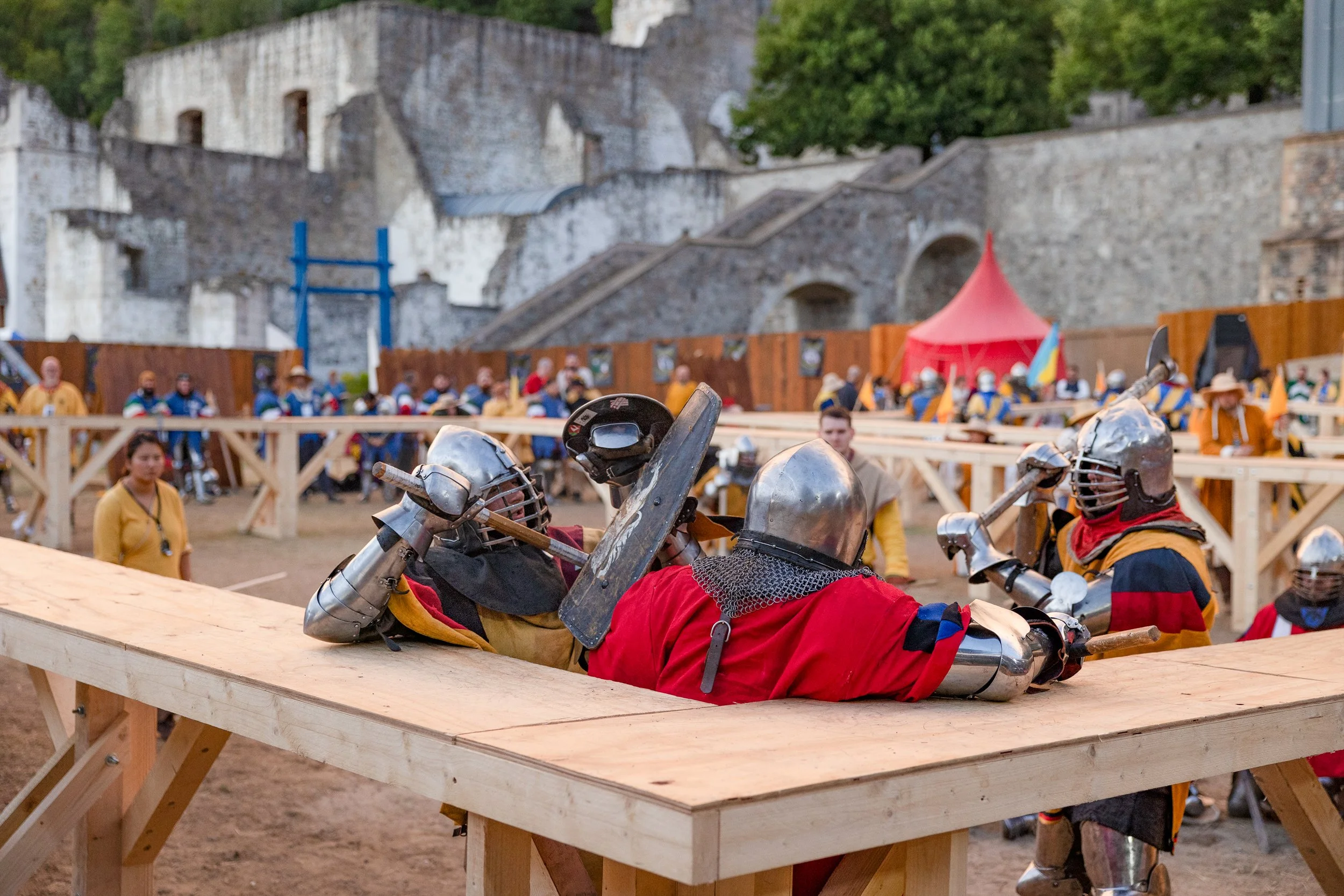 Reenactment of medieval knights engaged in combat, lying on a wooden platform with shields and weapons, in a historical setting with spectators and a castle in the background.