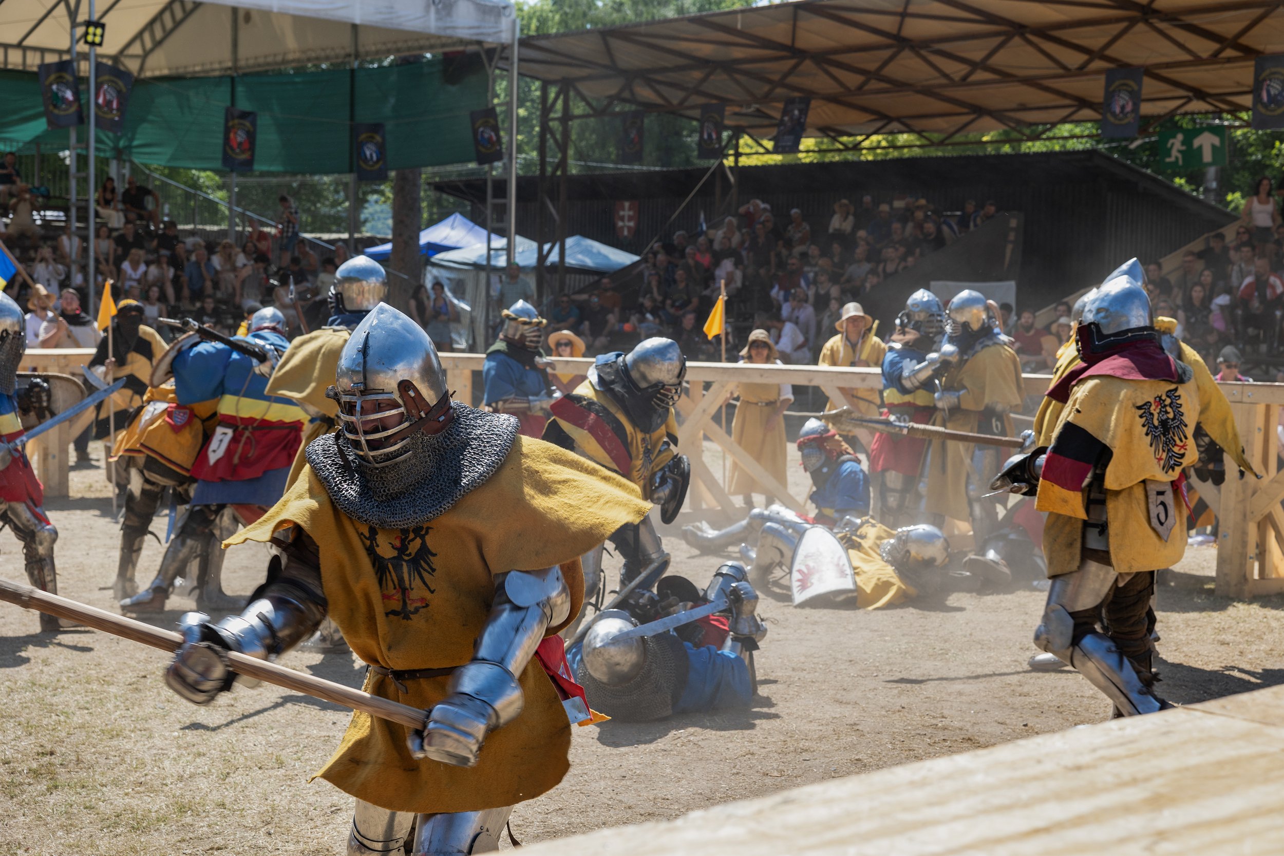 Medieval reenactors in armor and costumes participating in a historical sword fighting event at a fenced arena with spectators under a canopy in the background.
