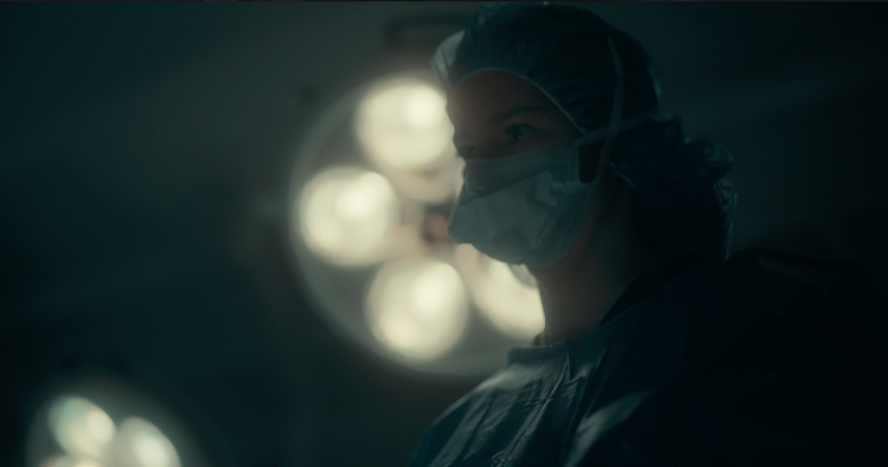 A female surgeon wearing a face mask, surgical cap, and gown looking to the side in an operating room with bright surgical lights in the background.