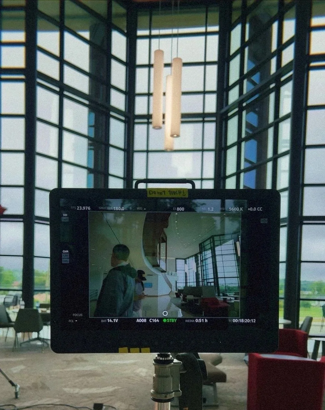 A camera monitor in a modern, spacious interior with large grid windows and hanging lights, capturing the scene of two people, one in a dark jacket and another in a cap, inside the building.