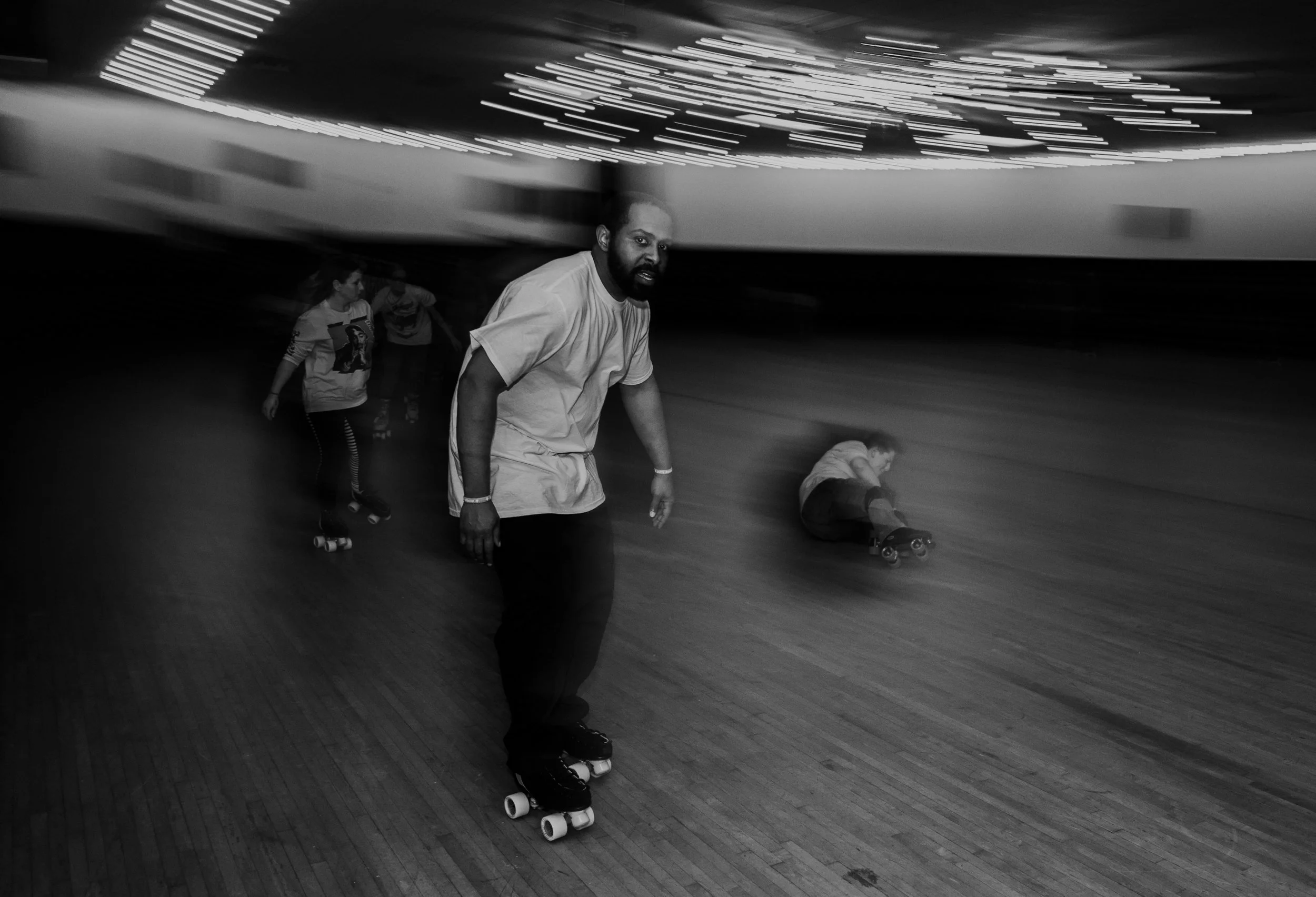 A man with a beard rollerskates on a wooden floor in an indoor skating rink, with children in the background, one sitting on the floor roller skating, black and white photo with motion blur.