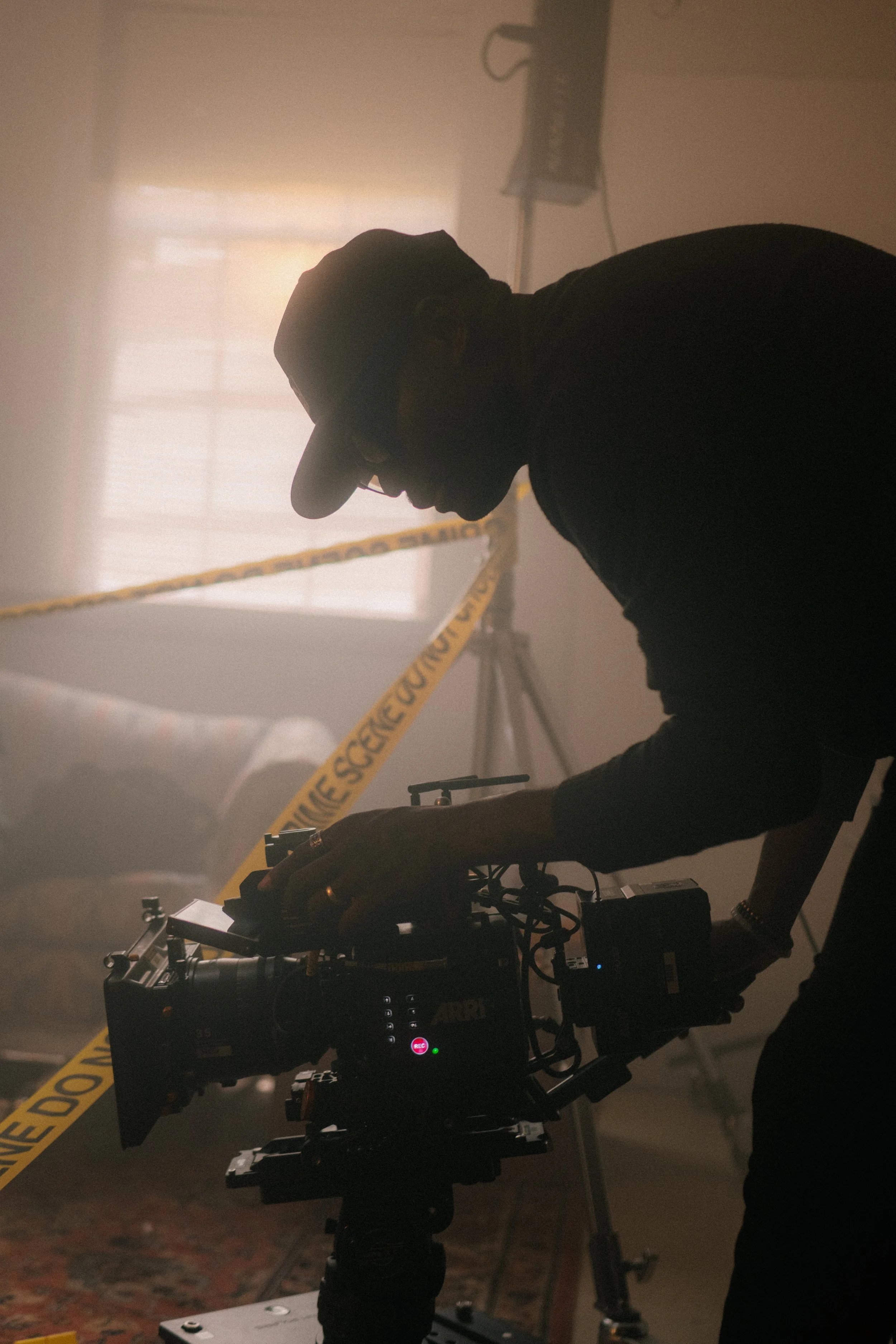 A person is operating a professional camera on a tripod in a dimly lit room with caution tape that reads 'crime scene do not cross' and a large window with blinds in the background.
