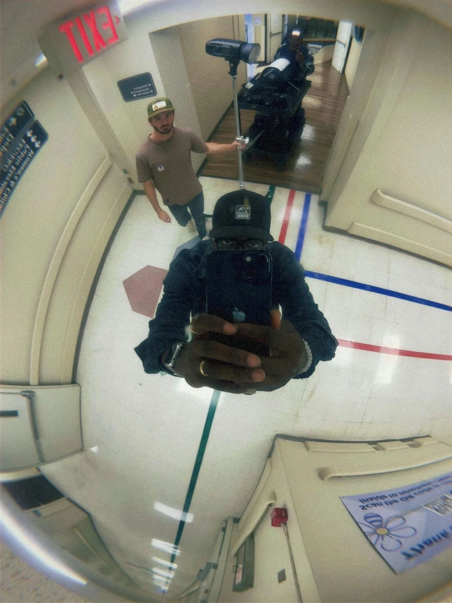 A person taking a selfie in a convex security mirror in a hallway, with another person standing nearby and luggage in the background