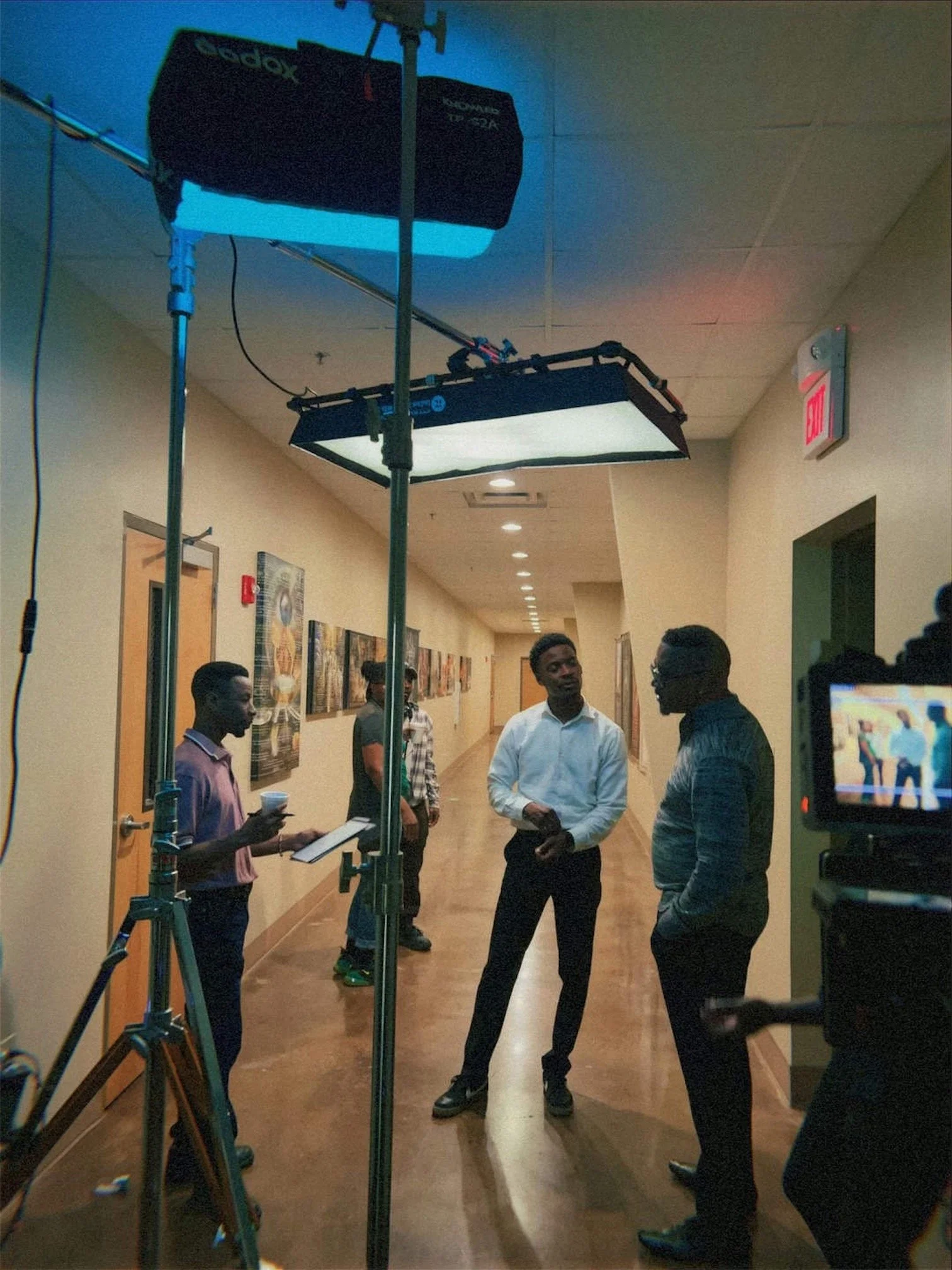 Group of people having a discussion in a hallway during a film or video shoot, with lighting equipment overhead.