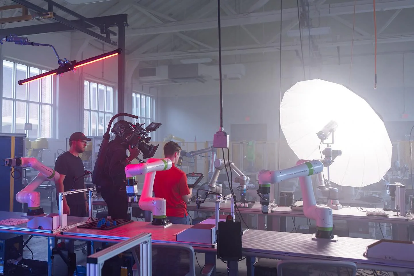 A robotics research lab with multiple robotic arms, a person operating a tablet, a camera on a moveable arm, and large studio lights, including a large white umbrella light.
