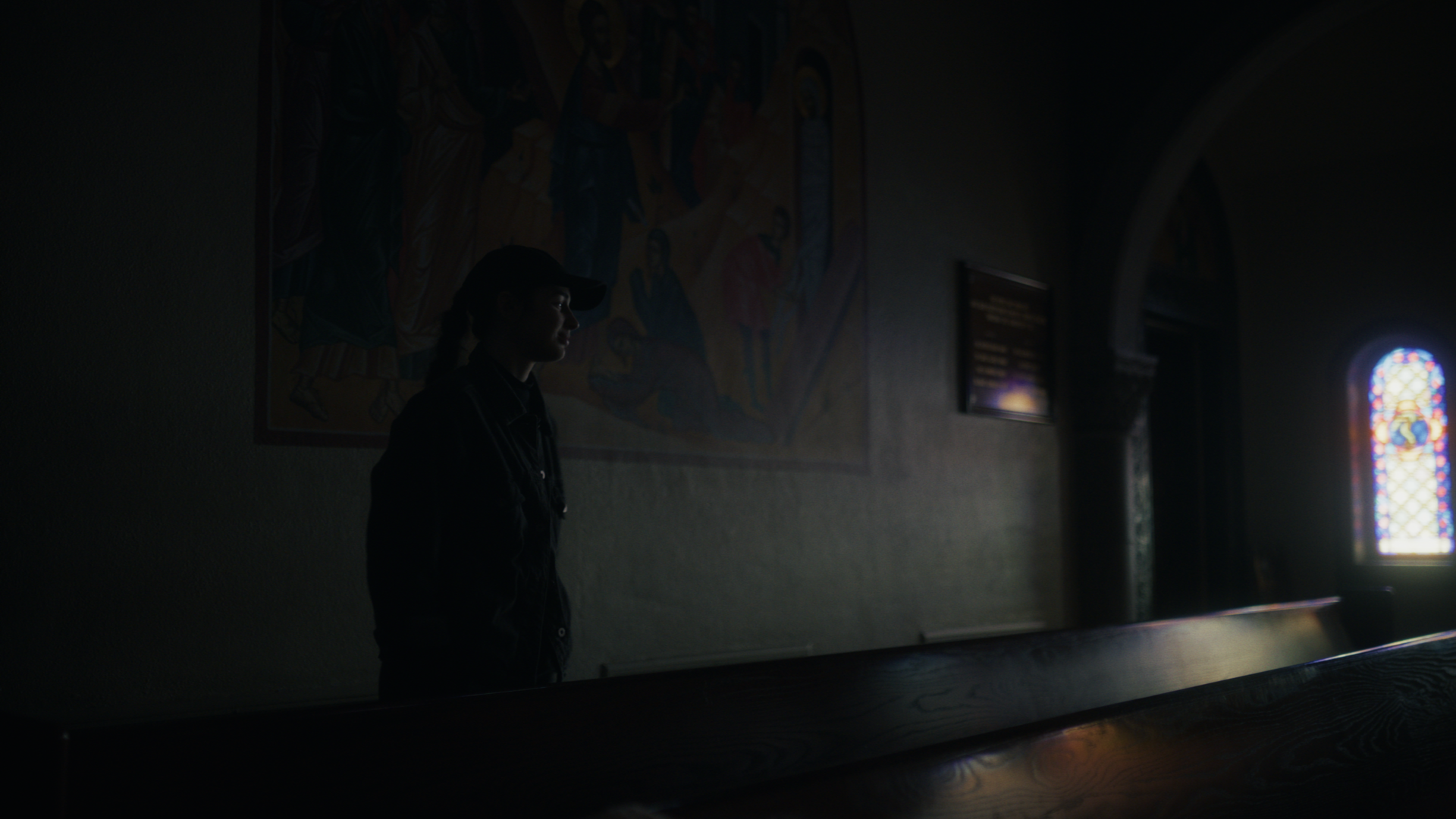 A woman in dark clothing and a baseball cap standing inside a dimly lit church near stained glass windows, with religious artwork on the wall.