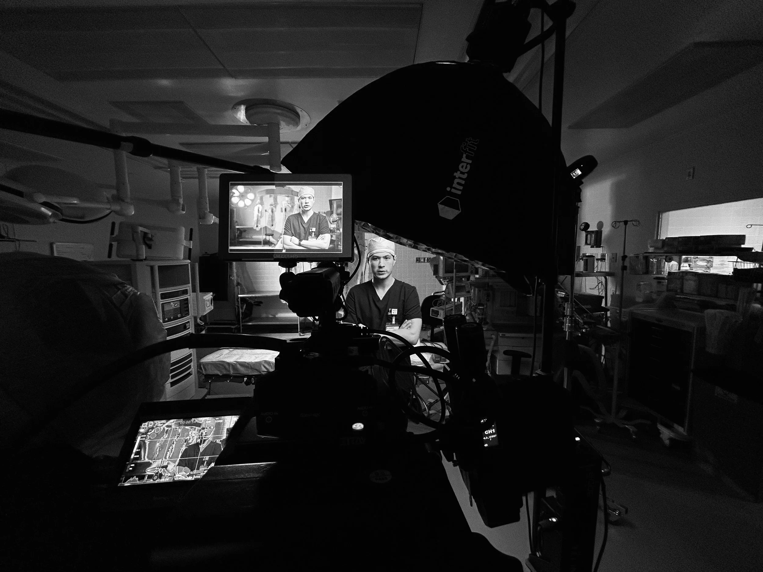 Filming equipment set up in a hospital room, with a person on camera and a monitor showing a man in medical scrubs.