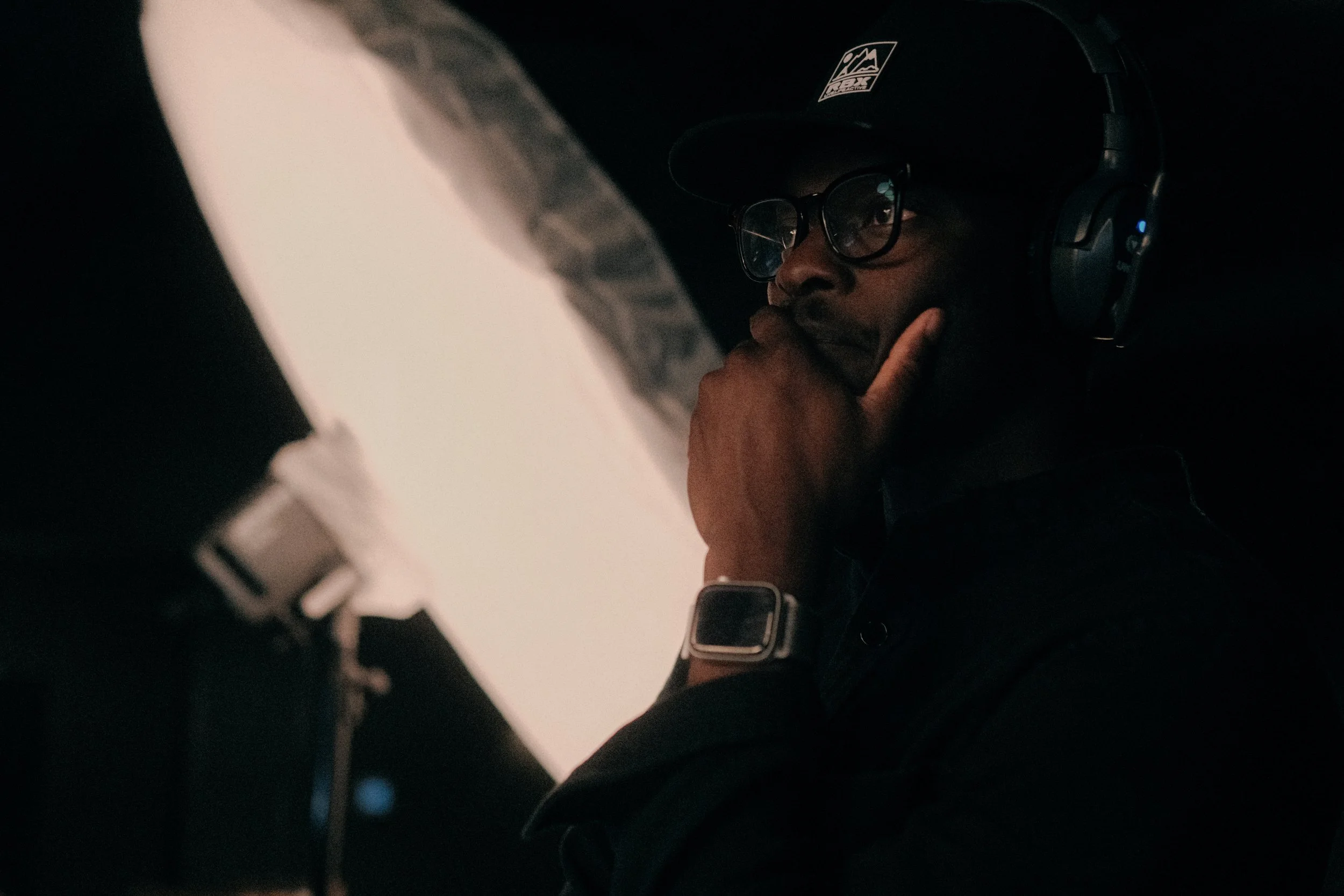 A man wearing glasses, a baseball cap, and headphones is sitting at a desk, looking at a computer screen with a pensive expression, hand on chin, in a dark room.
