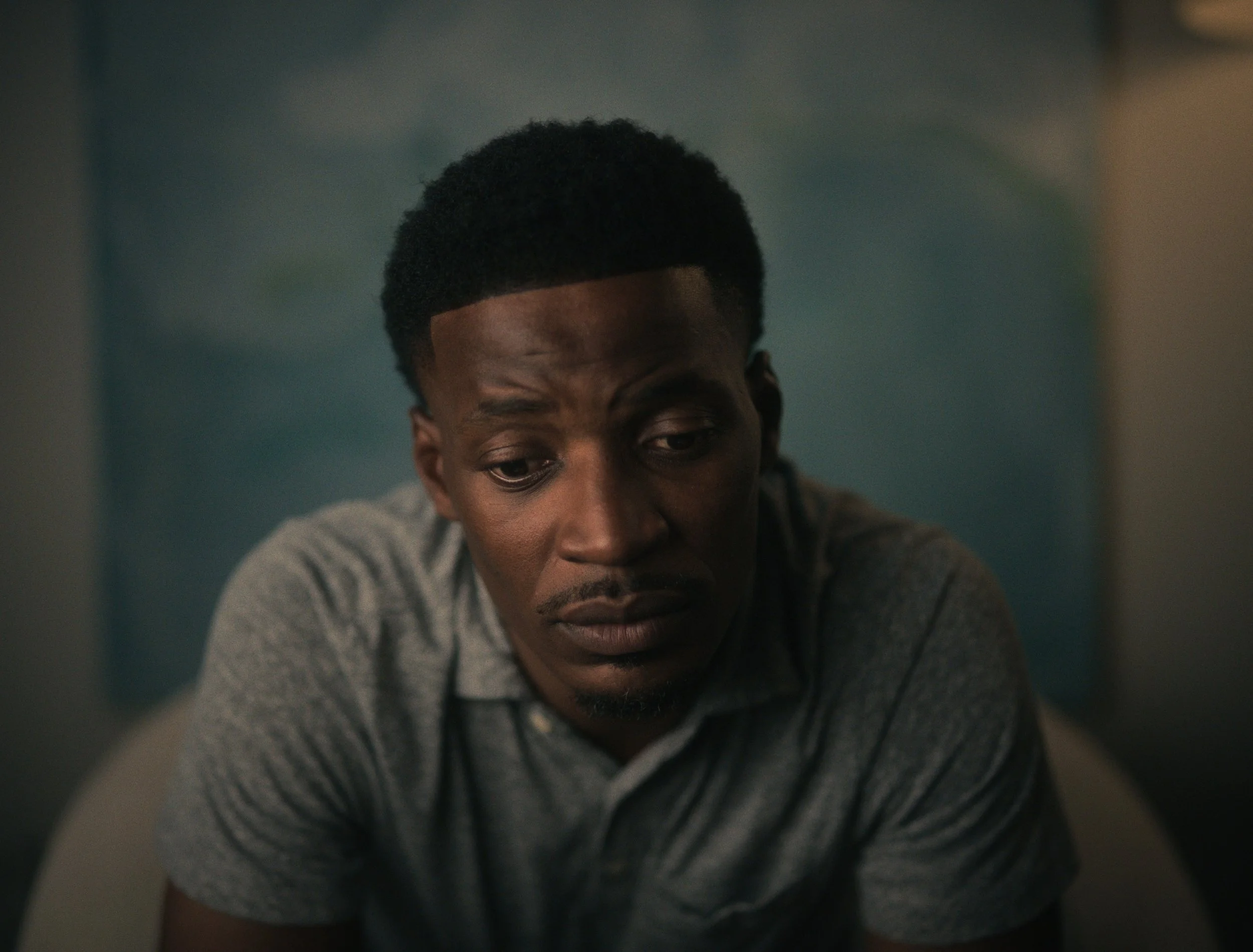 A man with short curly hair and a mustache looking down with a worried or contemplative expression, wearing a gray polo shirt, sitting on a chair in a room with blurred background.