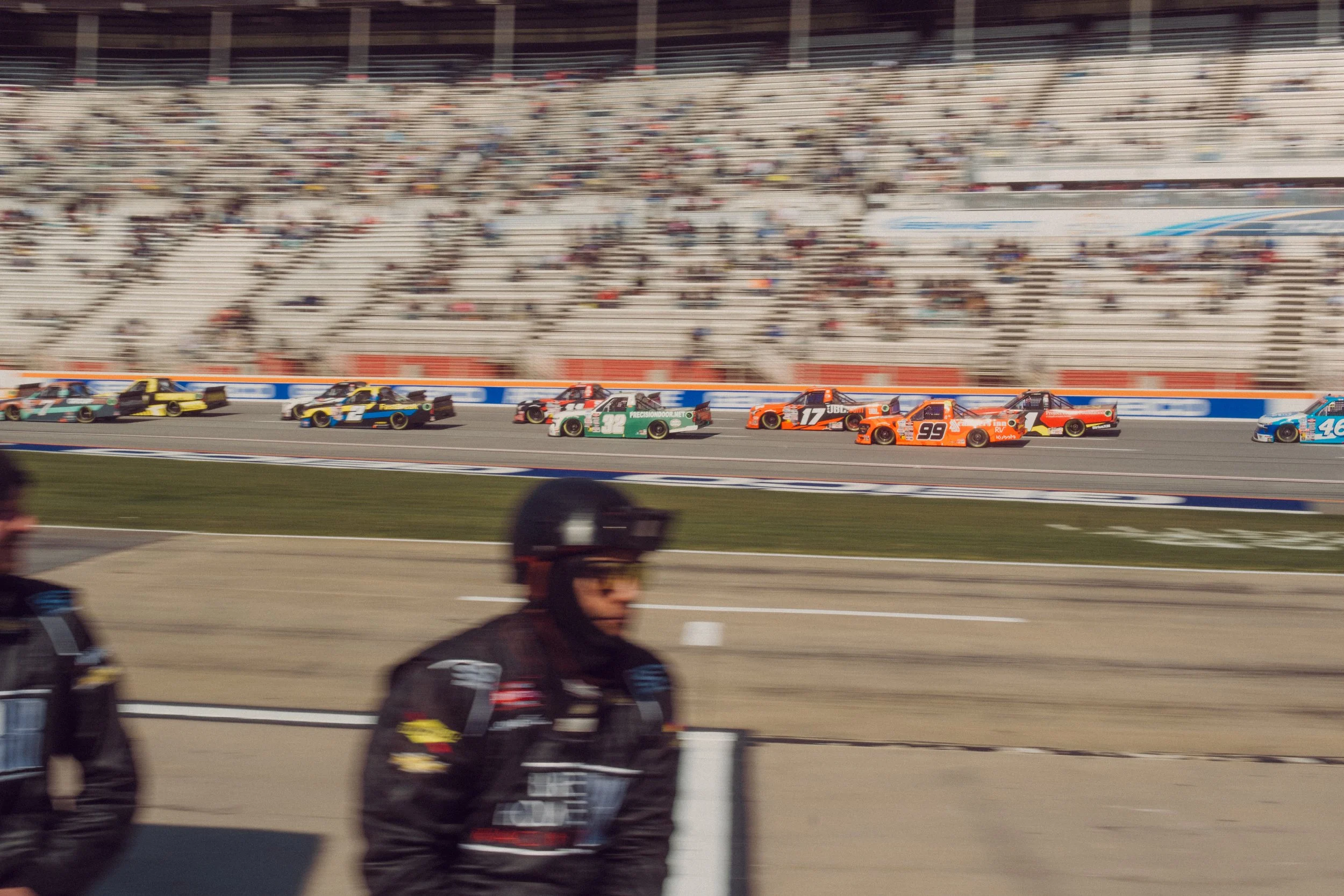 Race cars speeding on a racetrack with spectators in the stands and pit crew members in the foreground.