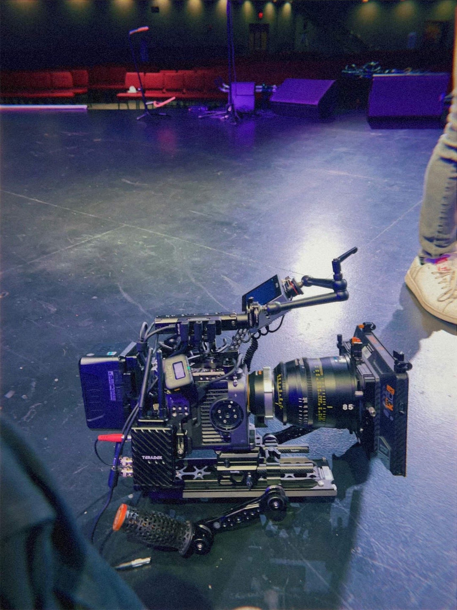 A professional camera on a camera rig on a dimly lit stage with some musical equipment and red seating area in the background.